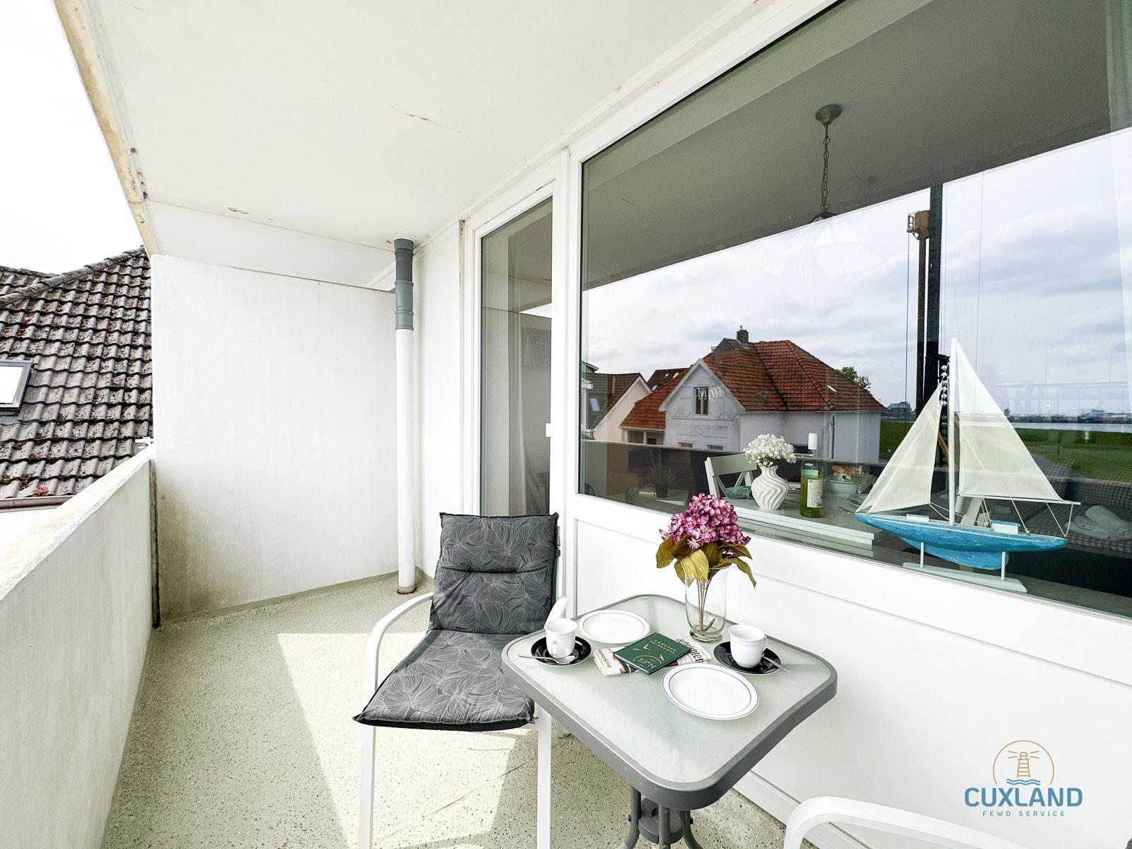Terrasse mit Tisch, Stuhl und Blumen. Durch Fenster ist Haus und Boot sichtbar.