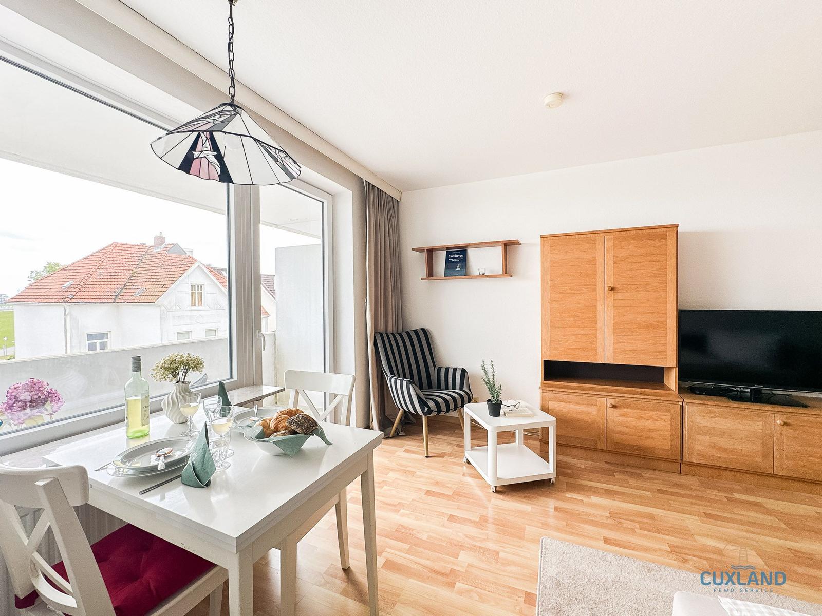 Essbereich mit Tisch, Stühlen und Blick auf Balkon. Wohnbereich mit TV, Schrank und Sessel.