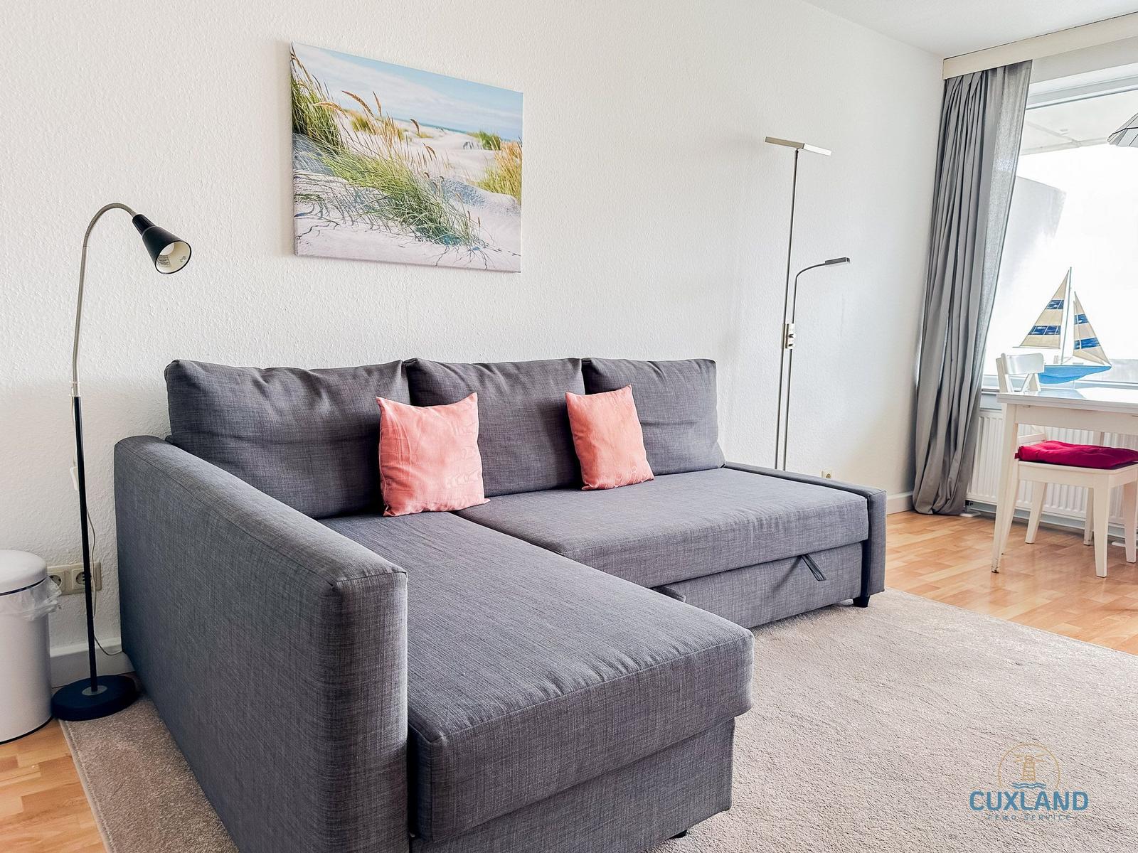 Ein grauer Sofa mit zwei rosa Kissen steht vor einer Wand mit Bild und Stehlampe.