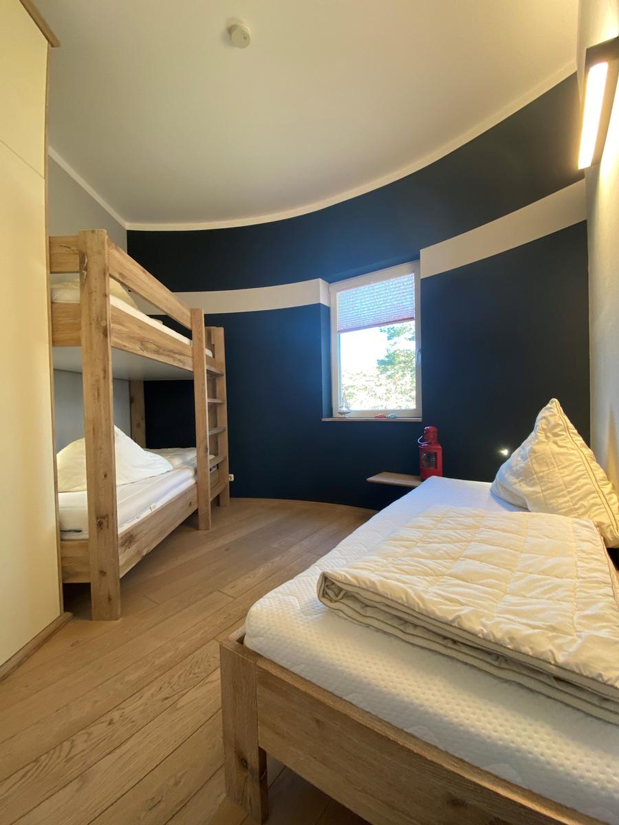 Zimmer mit Holzbett und Kojenbett, blauer Wand und Fenster mit Blick nach draußen.