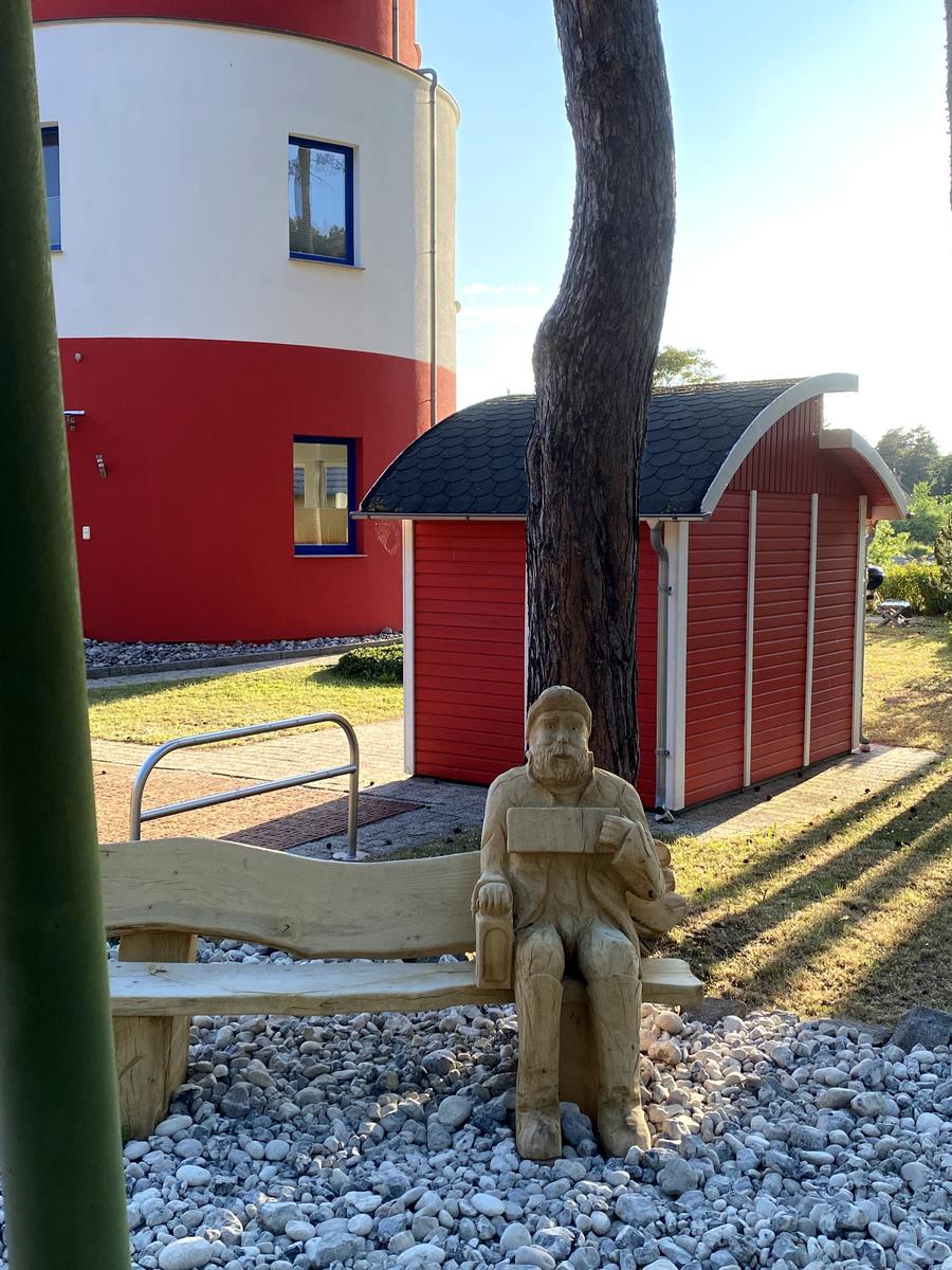 Holzsitzbank mit Skulptur vor rotem Gebäude und Baum.
