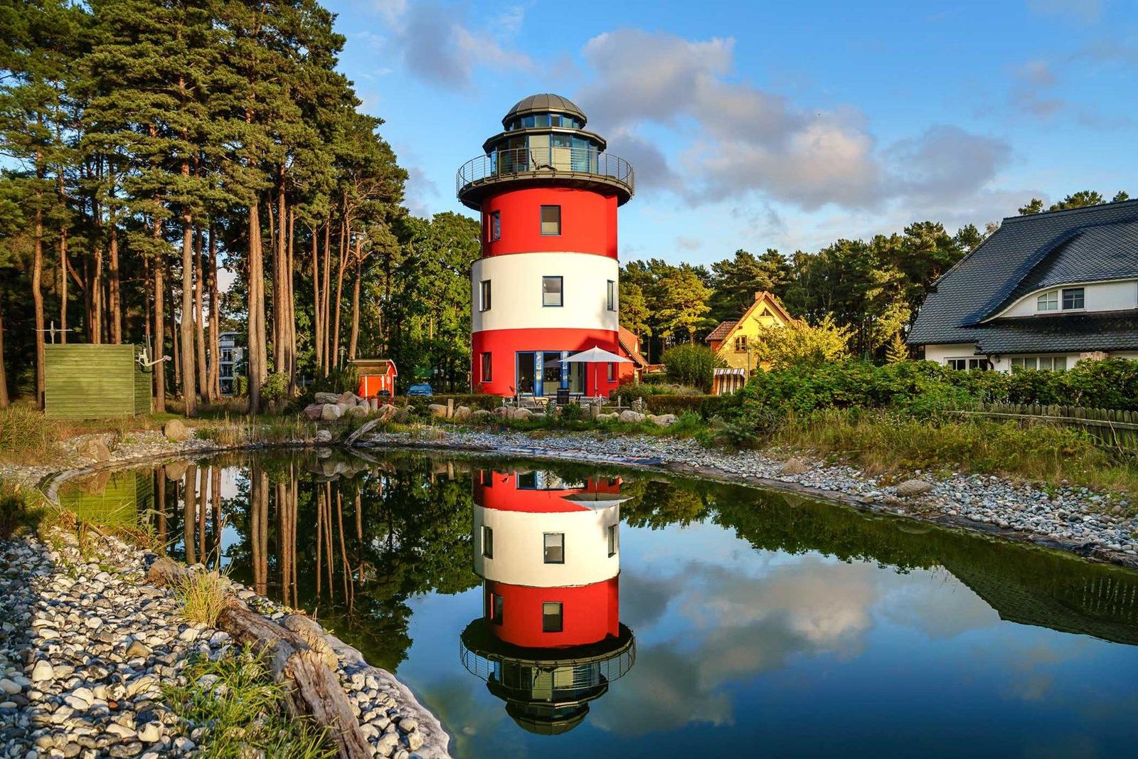 Rotes Leuchtturmhaus spiegelt sich im Wasser. Umgeben von Bäumen und Häusern.