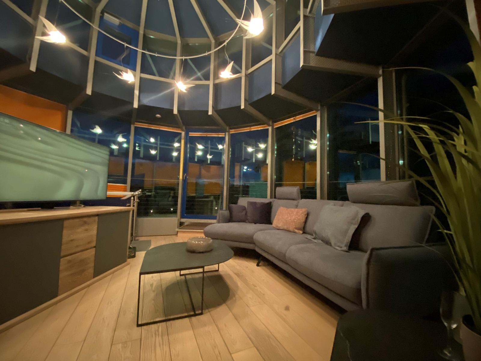 Wohnzimmer mit Sofa, Couchtisch und Fernseher. Große Fenster und Deckenleuchten.