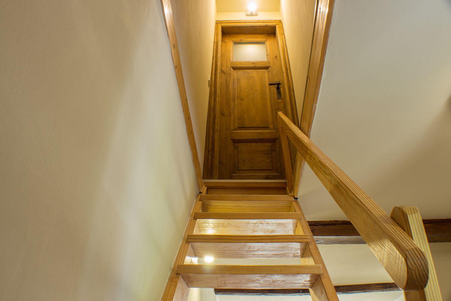 Holztreppe mit Handlauf führt zu einer Holztür. Deckenleuchte beleuchtet den Flur.