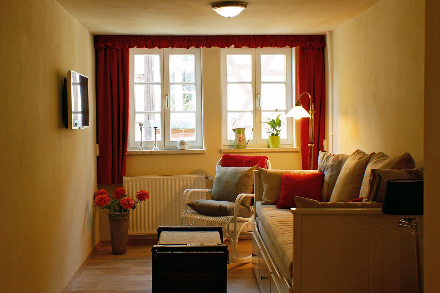 Wohnzimmer mit Sofa, Sessel, Fenstern, Vorhängen und Fernseher an der Wand.