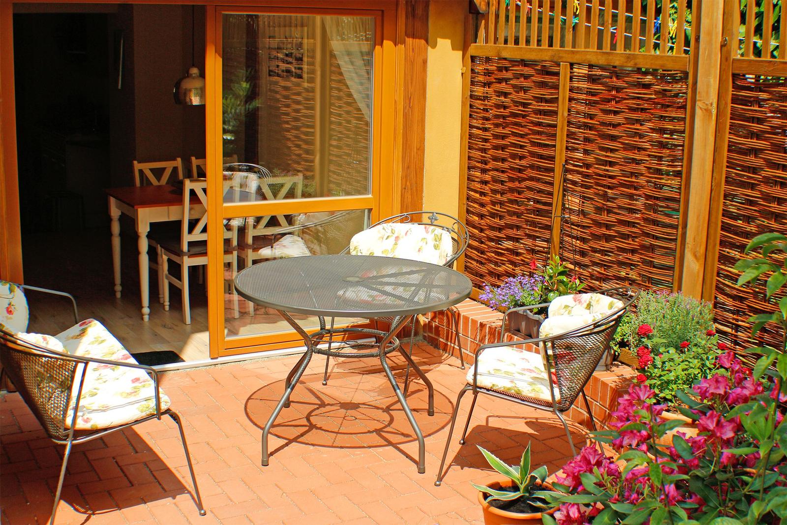 Terrasse mit runden Tisch und drei Stühlen, umgeben von Blumen und einem Holzzaun.