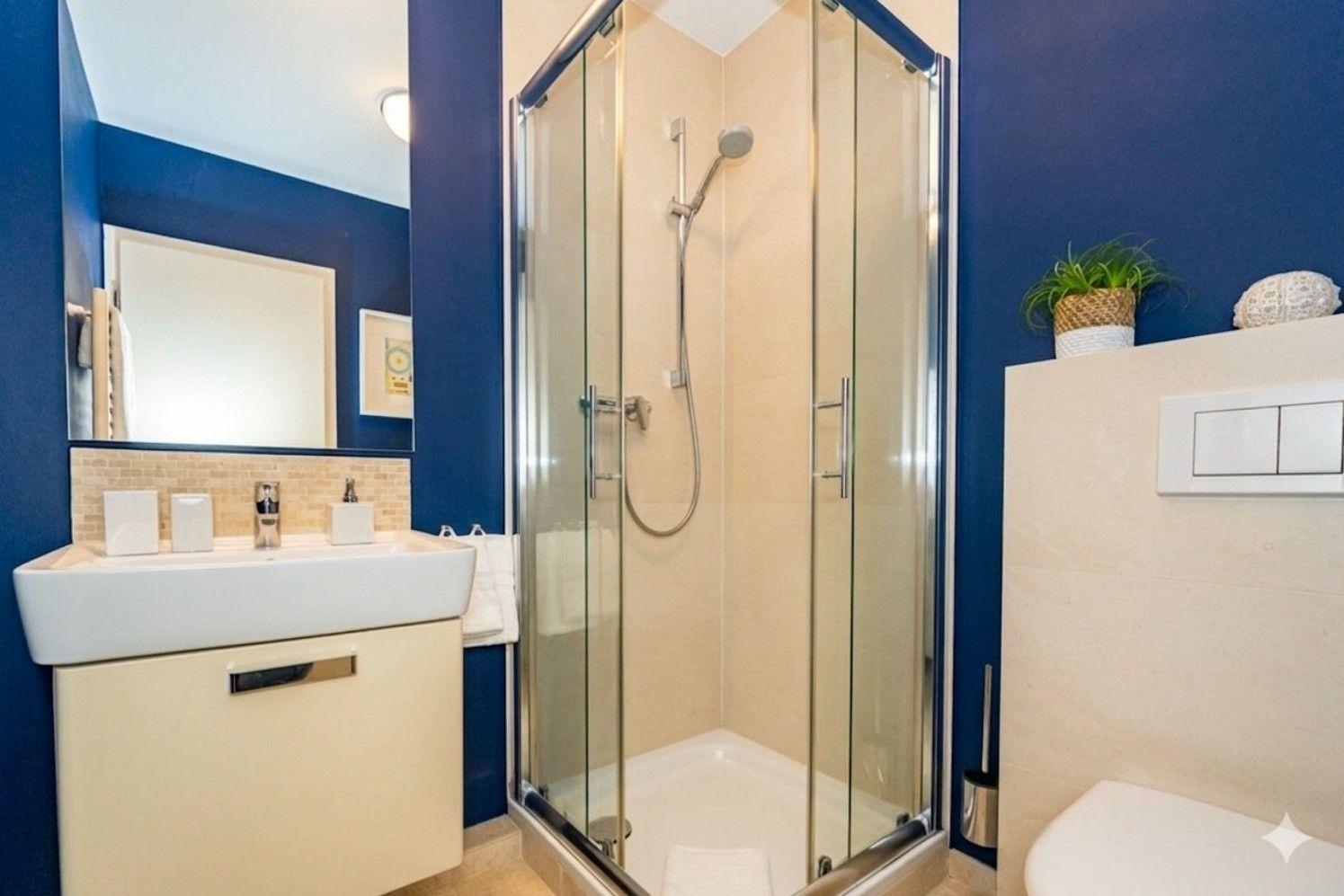 Badezimmer mit Dusche, Waschbecken und Toilette. Blaue Wände und helle Fliesen.