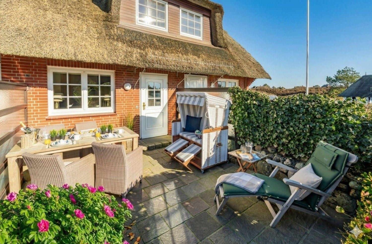 Terrasse mit Esstisch, Liegestühlen und Strandkorb vor einem strohgedeckten Haus.