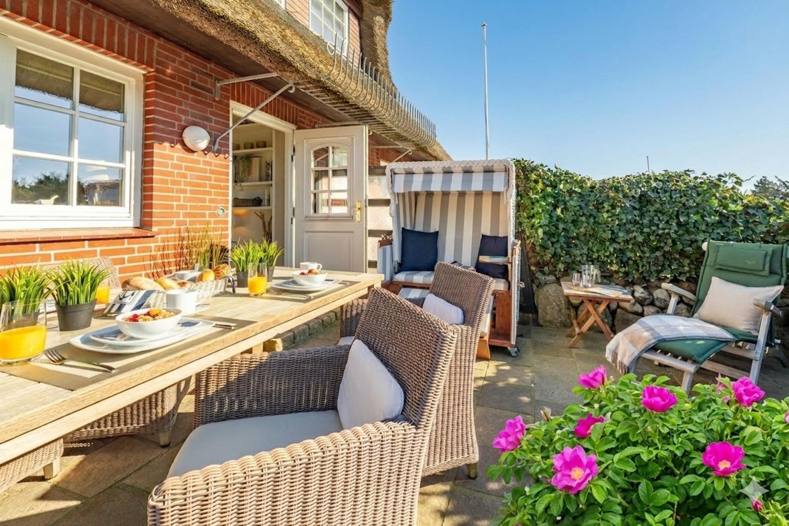 Terrasse mit Esstisch, Stühlen und Strandkörbe. Rosenblüten und grüne Hecke im Vordergrund.