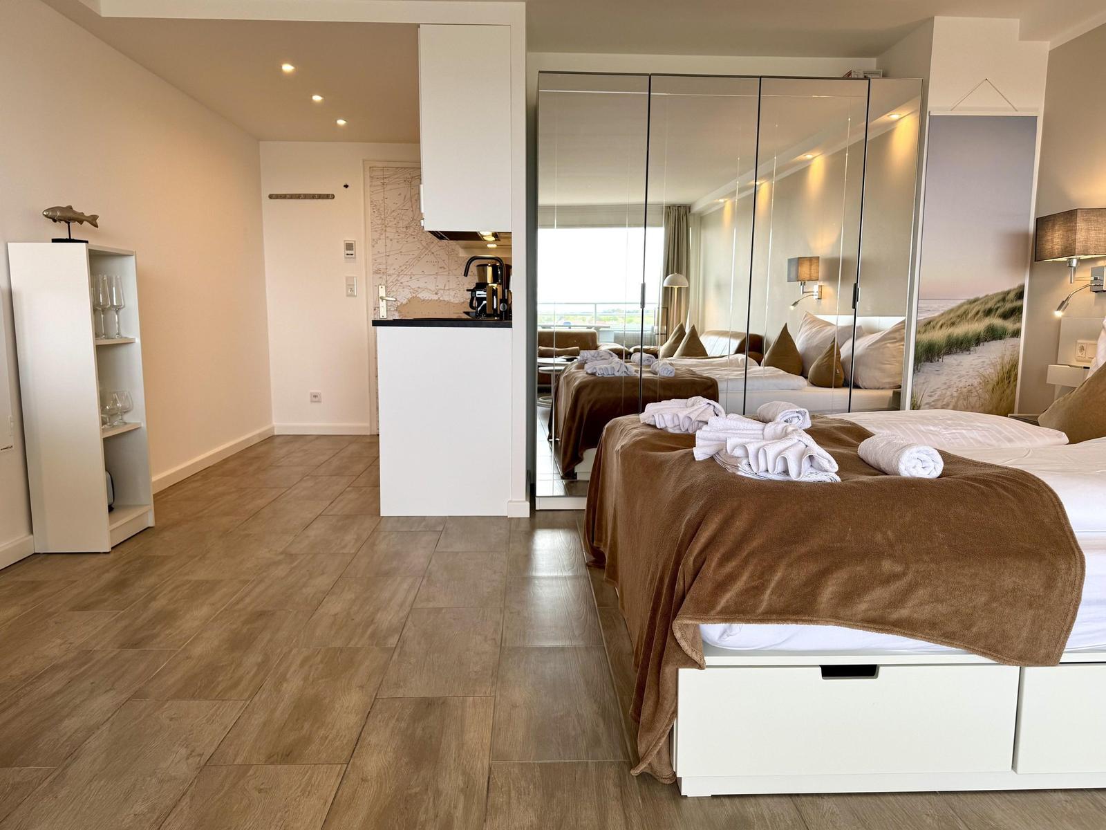 Schlafzimmer mit Bett, Spiegelschrank und Küchenbereich im Hintergrund.