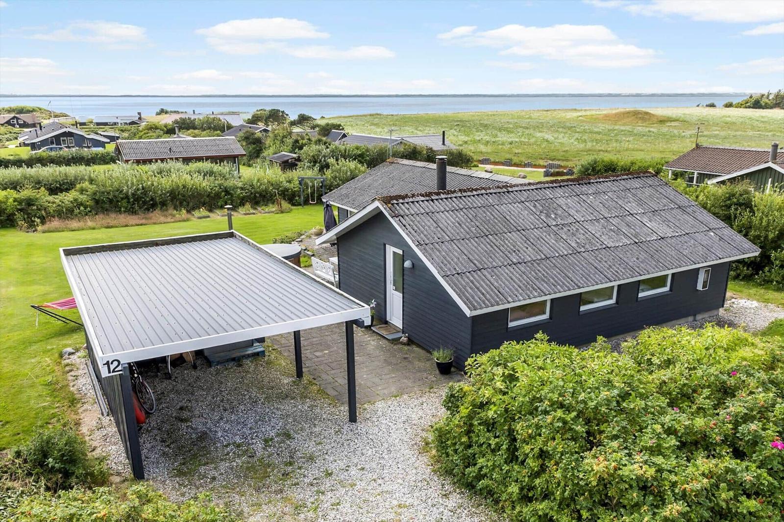 Ein graues Haus mit Carport und Garten, Blick aufs Wasser.