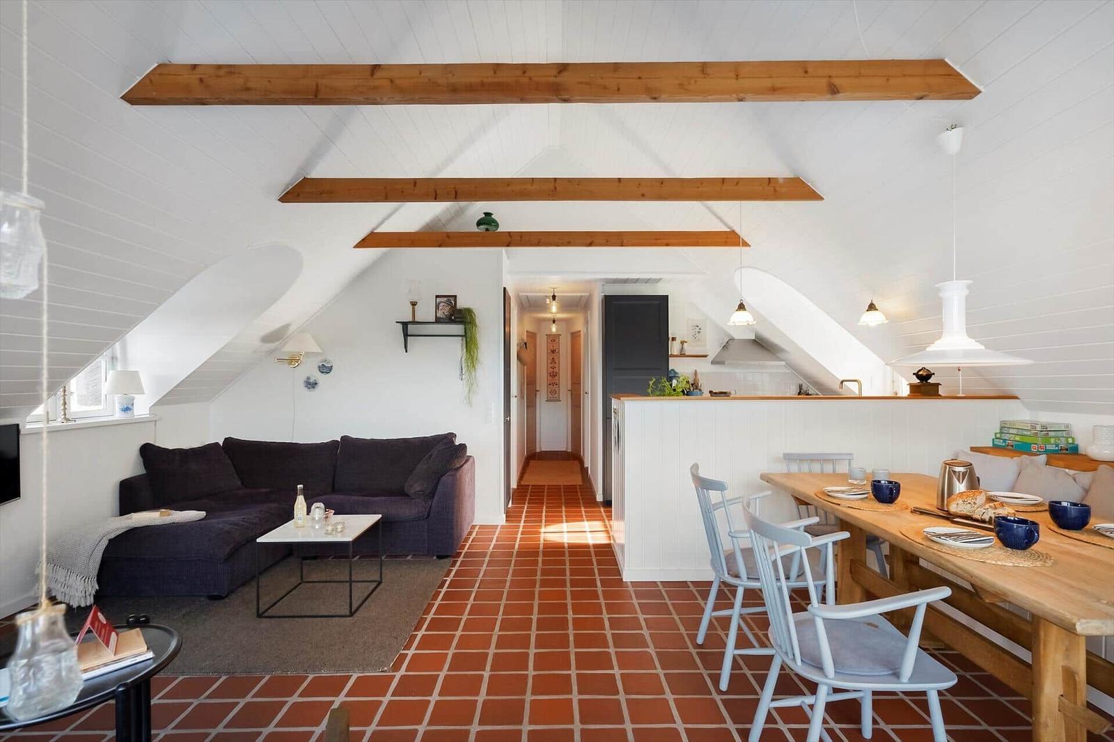 Wohn- und Essbereich mit Holzbalken, rotem Fliesenboden und dunklem Sofa.