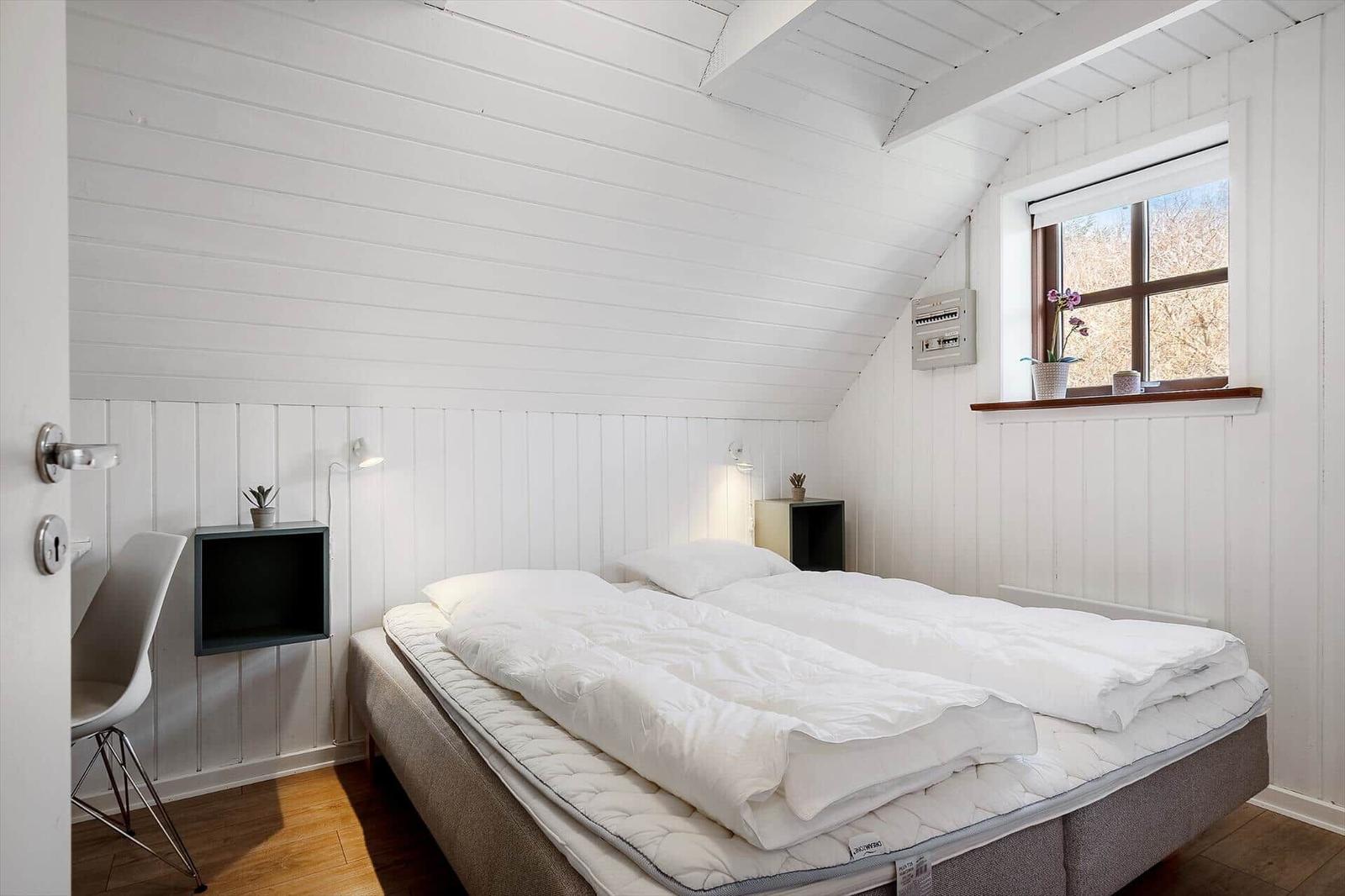 Schlafzimmer mit Doppelbett, weißer Wandverkleidung und Fenster mit Pflanze.