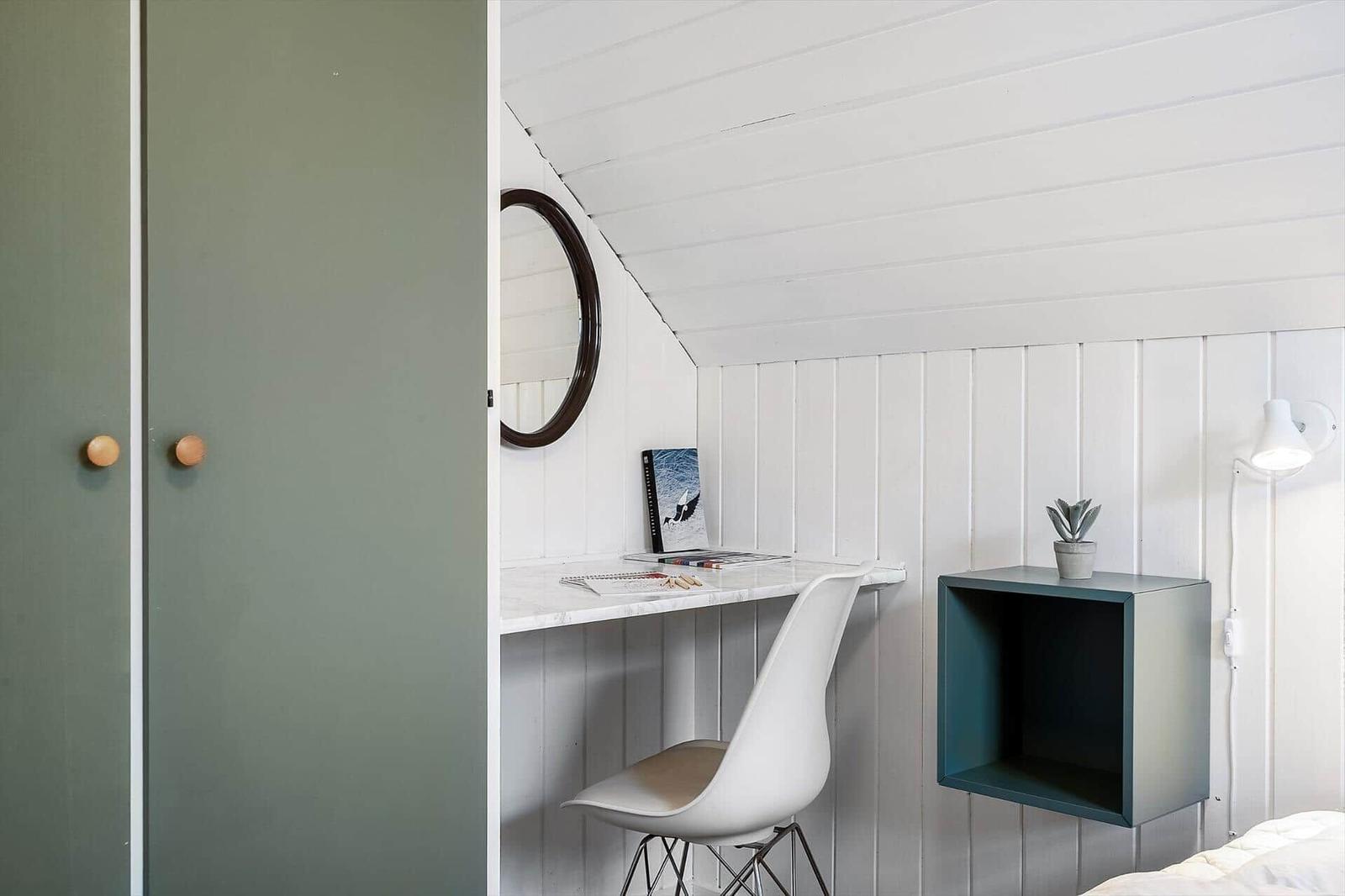 Schreibtisch mit Stuhl, Spiegel und Pflanze an weißer Wand. Grüner Schrank mit Holzgriffen.