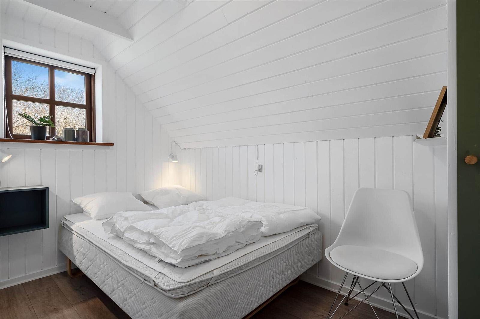 Schlafzimmer mit weißem Bett, Stuhl und Fenster. Wände und Decke weiß verkleidet.