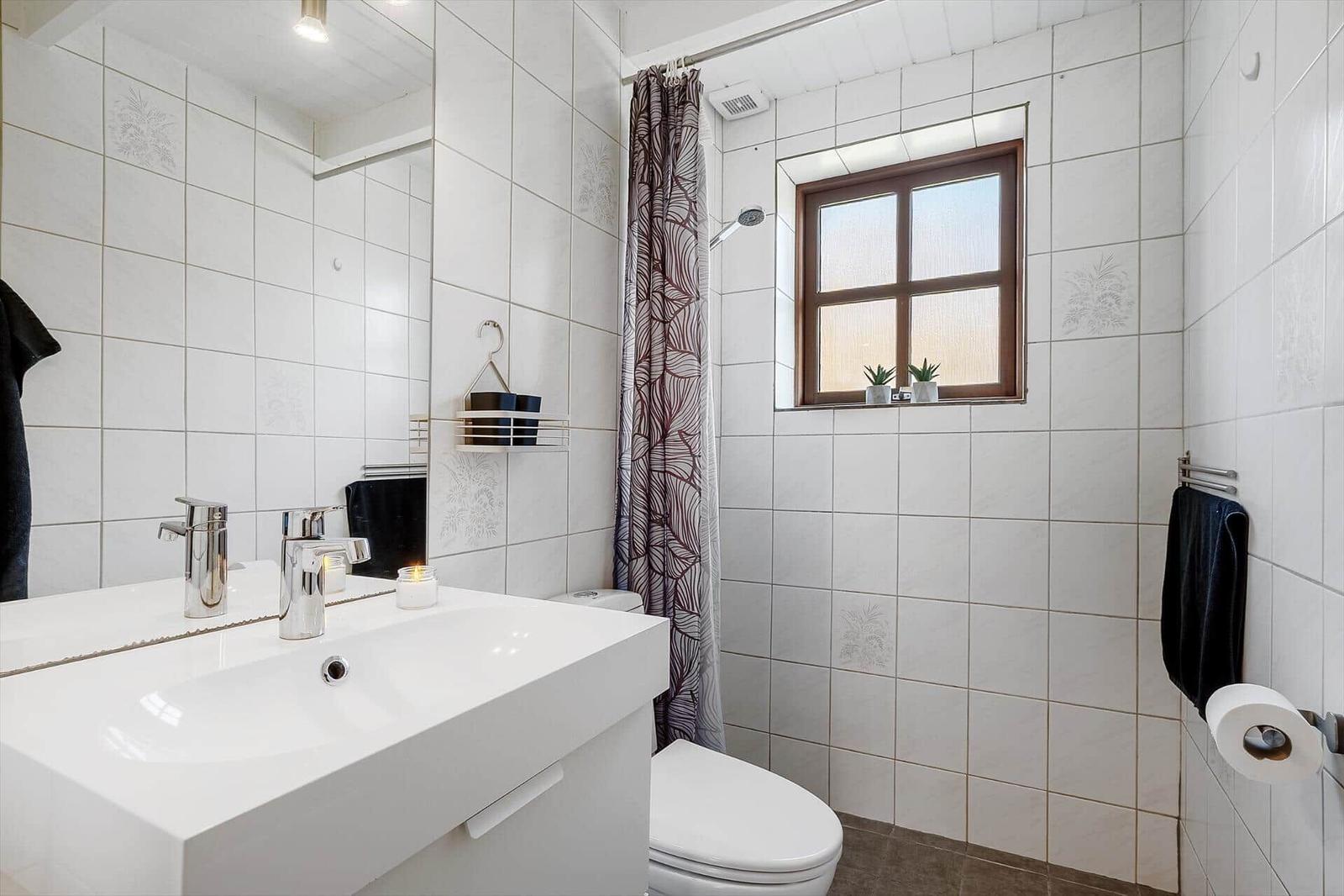Badezimmer mit Waschbecken, Toilette und Dusche. Fenster mit Pflanzen und Vorhang.