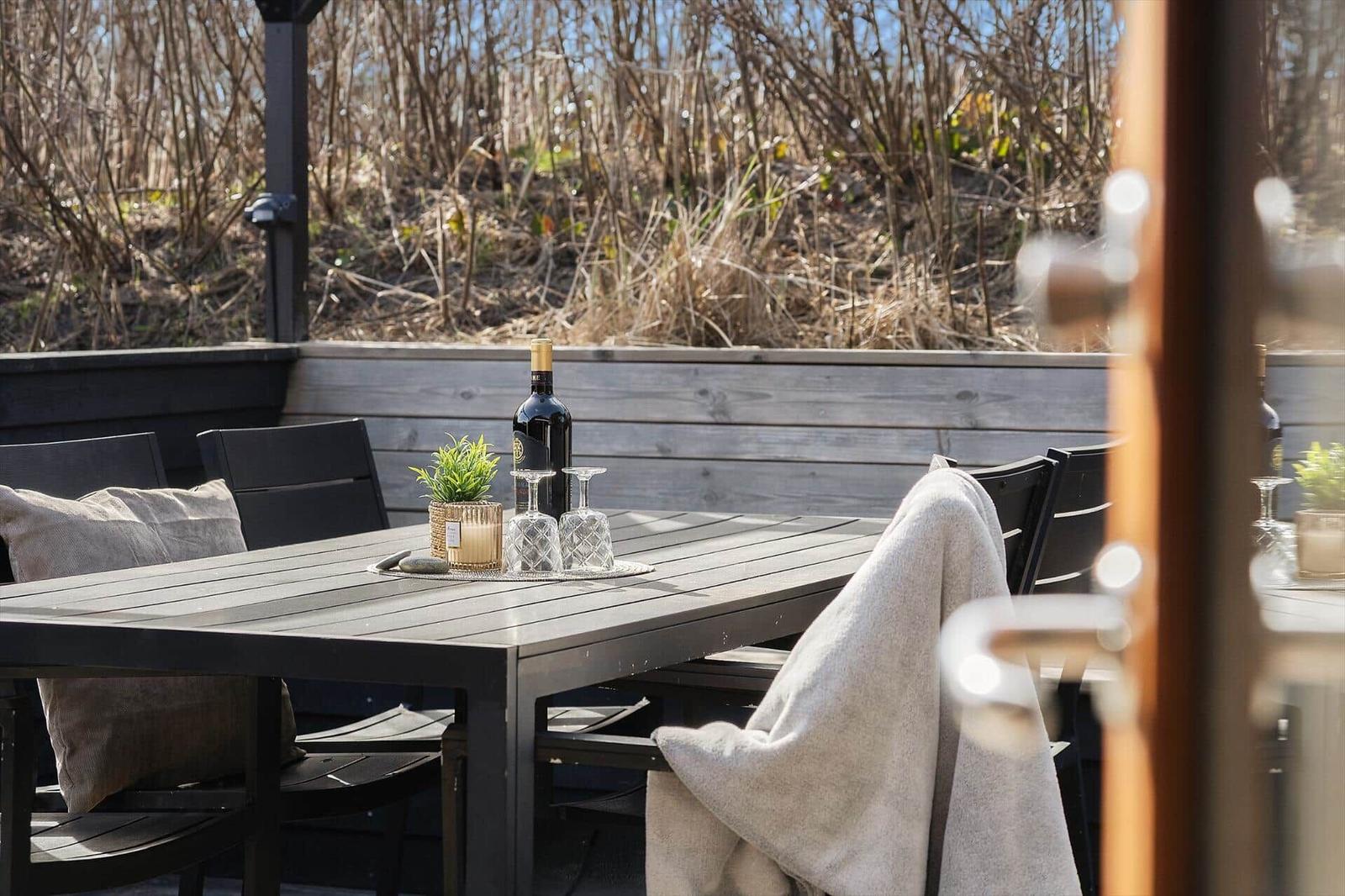 Terrasse mit Tisch, Stühlen und einer Weinflasche. Hintergrund: trockenes Gras.