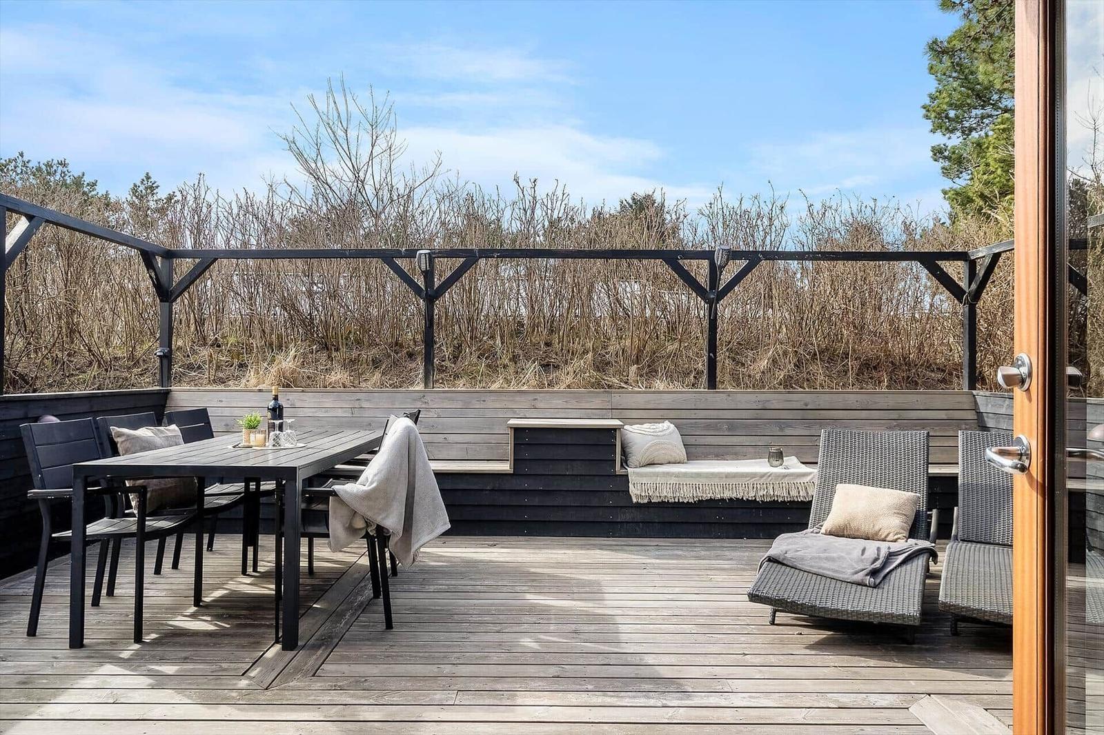 Terrasse mit Tisch, Stühlen und Liegen. Hintergrund: Hecken und Himmel.