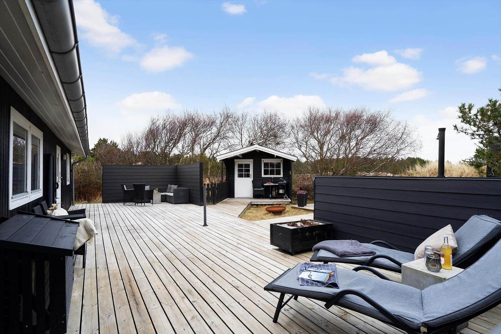 Großer Holzterrasse mit Liegen, Grill und Sitzgelegenheiten. Blick auf kleine Hütte und Bäume.