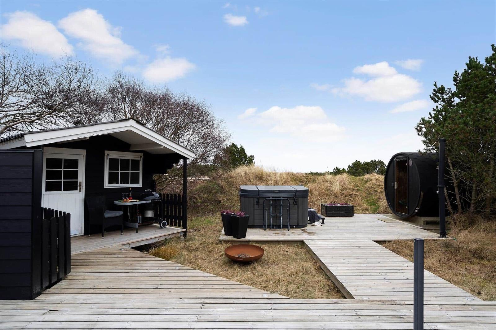 Holzterrasse mit Whirlpool, Sauna und Grillplatz vor einem schwarzen Holzhaus.