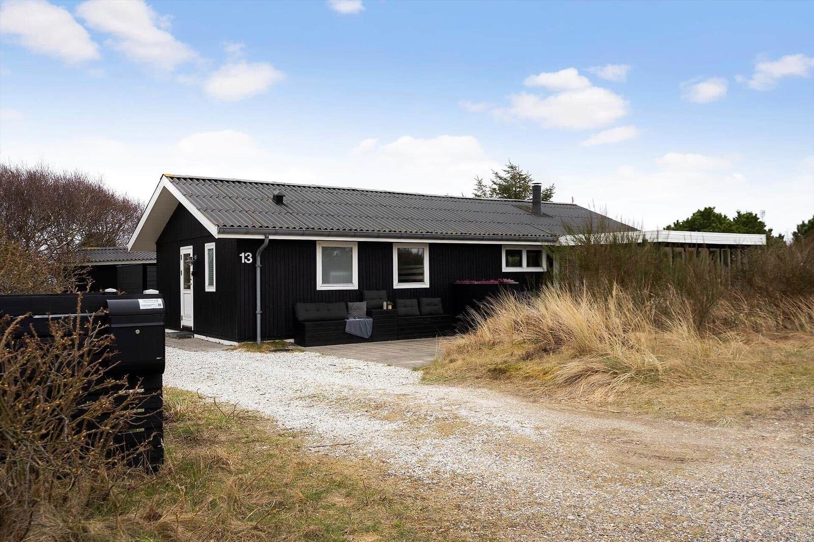 Schwarzes Ferienhaus mit Terrasse und Garten. Hausnummer 13. Gravelweg und trockenes Gras.