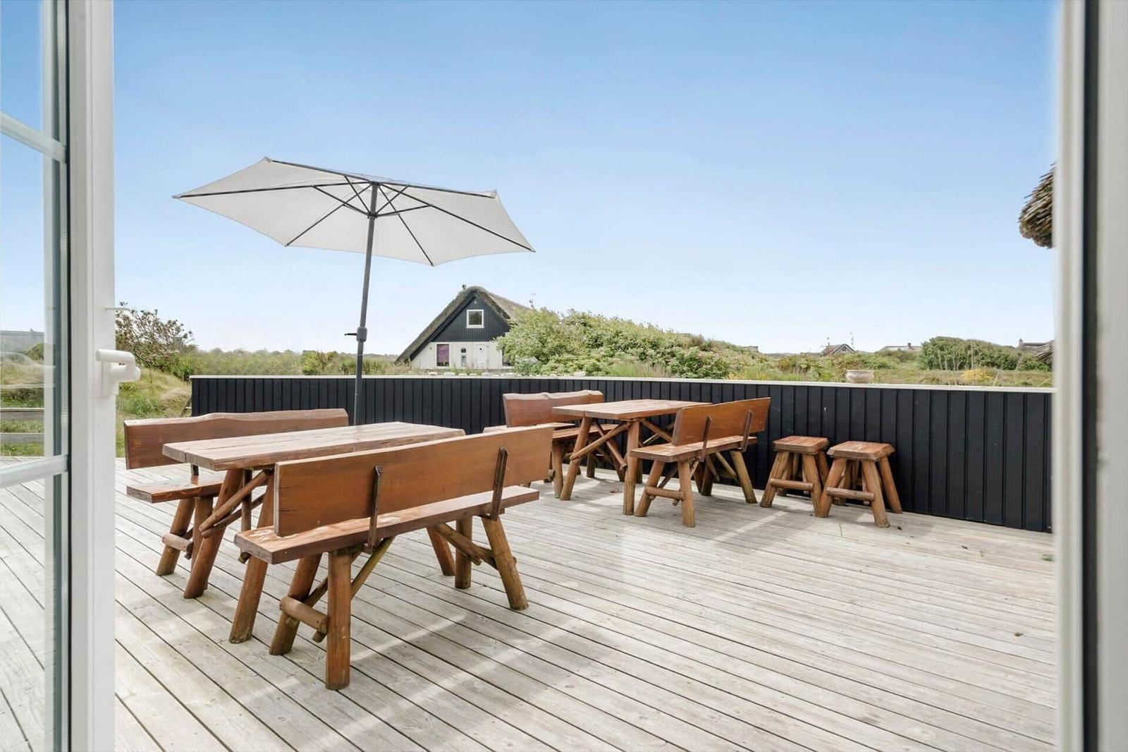 Terrasse mit Holztischen, Bänken und Sonnenschirm. Blick auf grüne Landschaft und Haus.