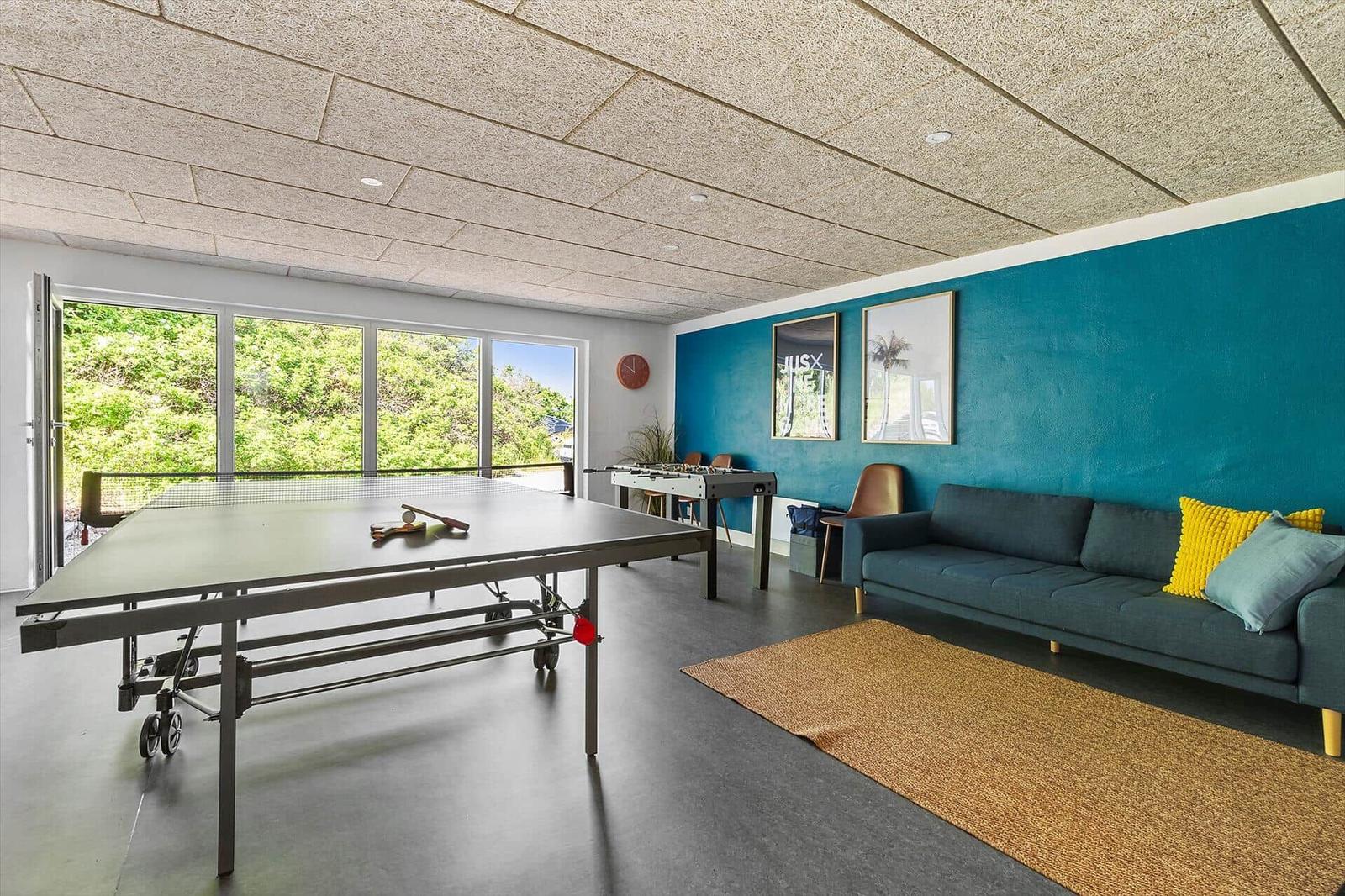 Raum mit Tischtennisplatte, Sofa und Foosballtisch. Blaue Wand und große Fenster.