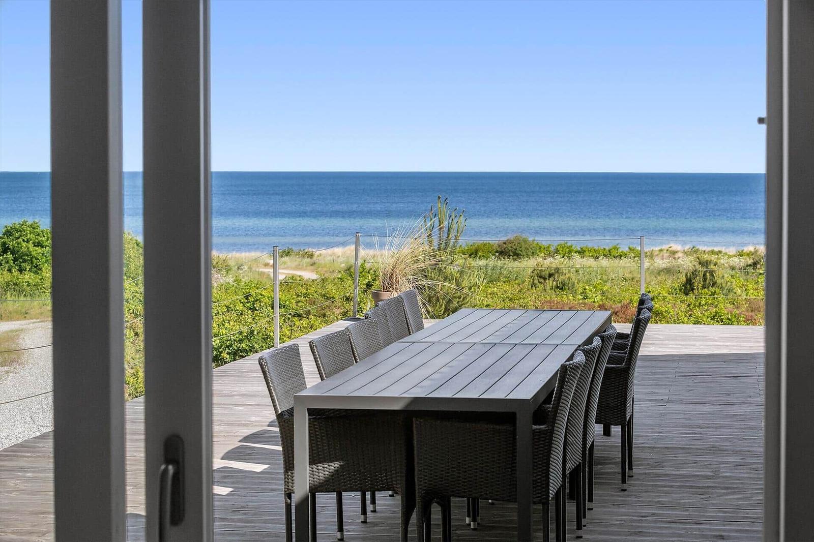 Terrasse mit Tisch und Stühlen bietet Meerblick. Holzdeck und Grünflächen sichtbar.