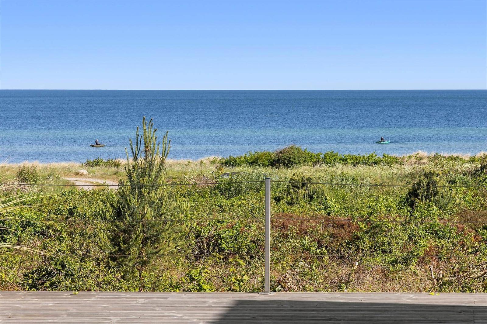 Holzterrasse mit Blick aufs Meer und Strand. Zwei Personen auf dem Wasser.
