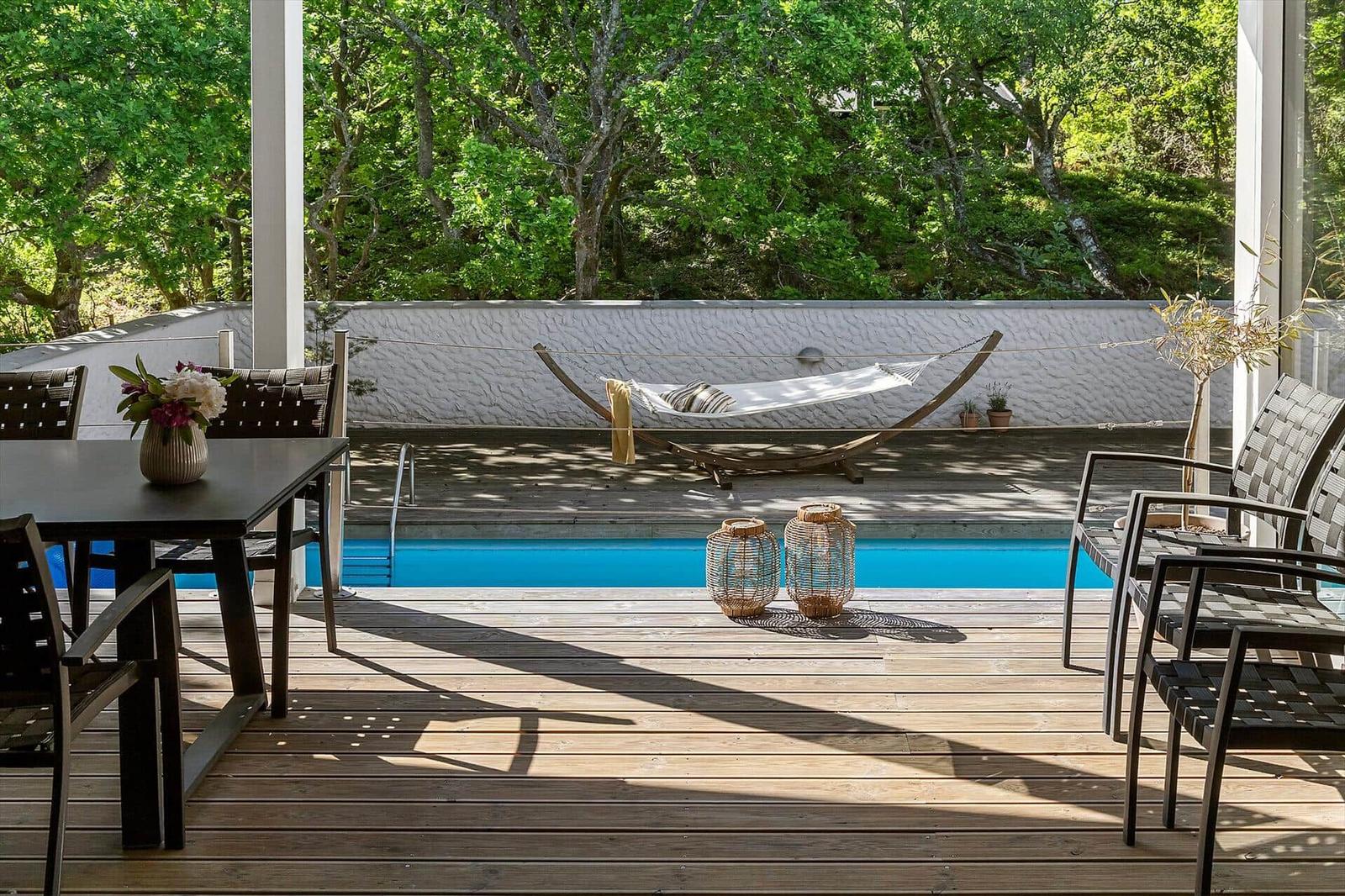 Poolbereich mit Liege, Tisch und Stühlen auf Holzterrasse umgeben von Bäumen.