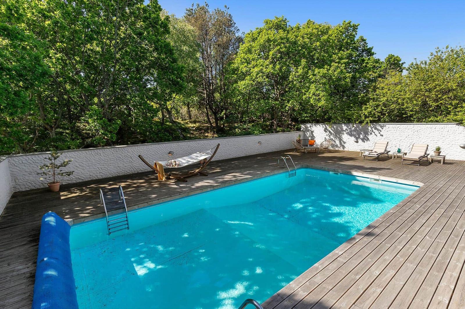 Pool mit Holzterrasse, Liegestühle und Hängematte umgeben von Bäumen.