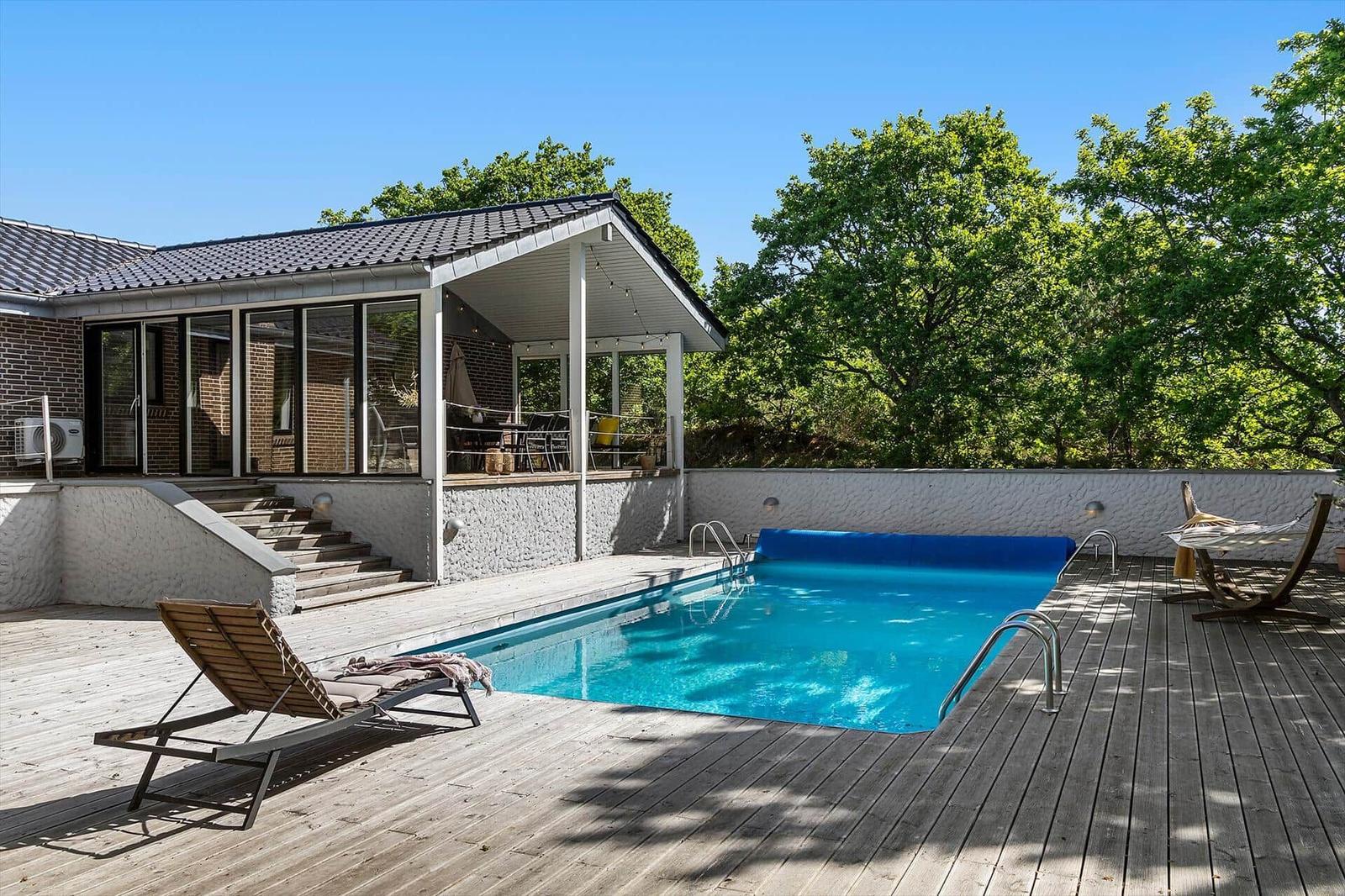 Pool mit Holzterrasse, Sonnenliege und Blick auf grüne Bäume.