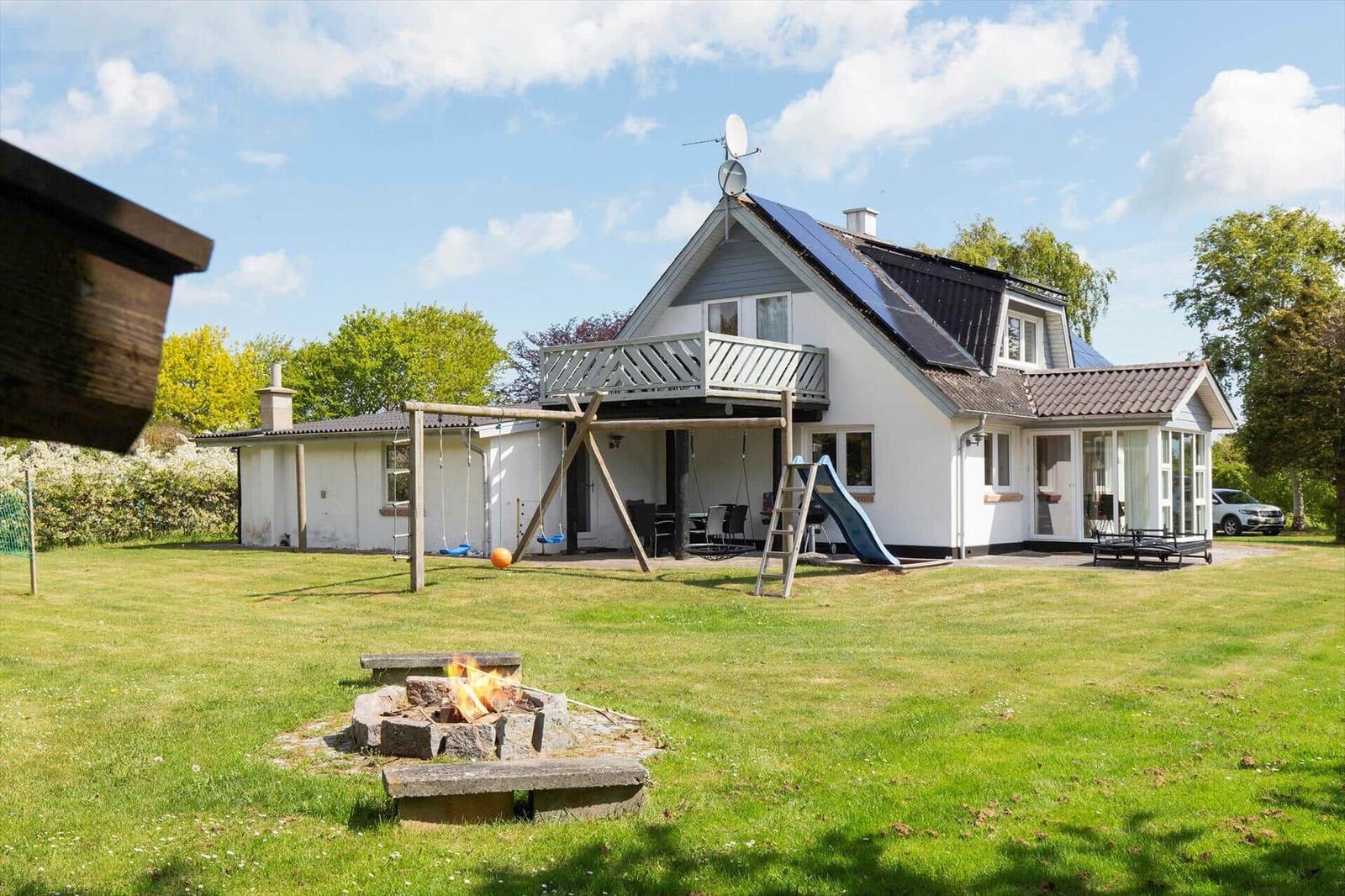 Haus mit Spielplatz, Grillbereich und Garten. Ein Feuer brennt im Steinbecken.