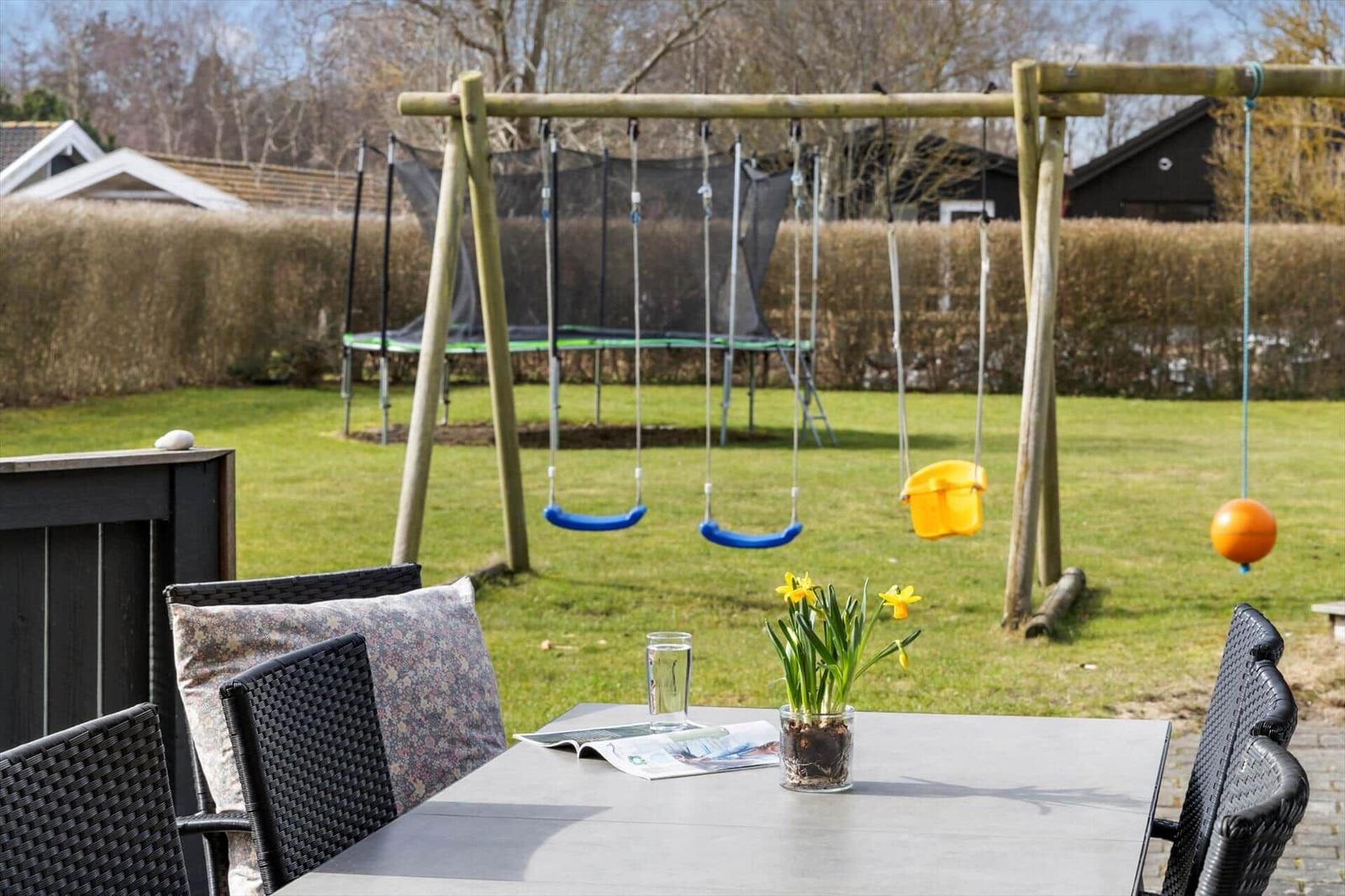 Terrasse mit Tisch, Stühlen und Blumen. Hintergrund: Spielplatz mit Schaukel und Trampolin.