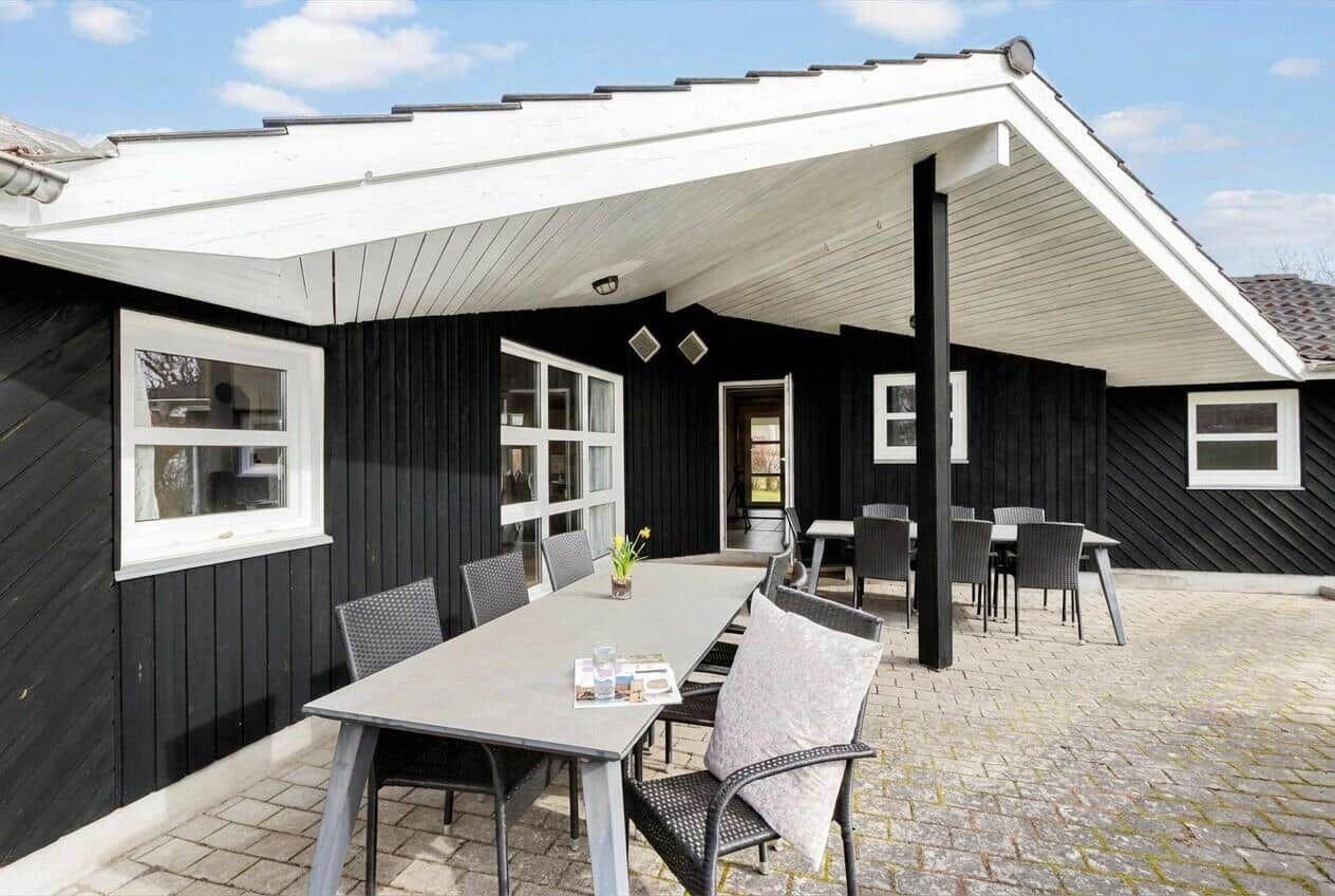 Terrasse mit Tisch und Stühlen unter Dach. Schwarze Fassade, weiße Fenster und Pflastersteinboden.