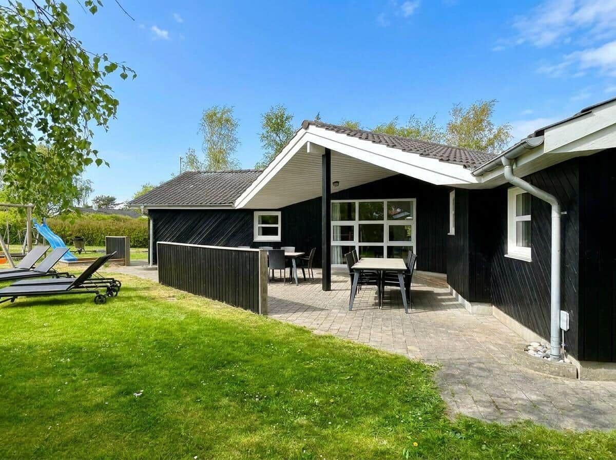 Schwarzes Holzhaus mit Terrasse, Garten und Liegestühlen unter blauem Himmel.