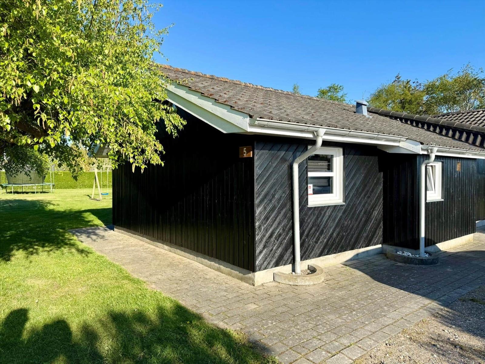 Schwarzes Holzhaus mit Dachrinne, Fenstern und Zufahrt. Grasfläche und Baum sichtbar.
