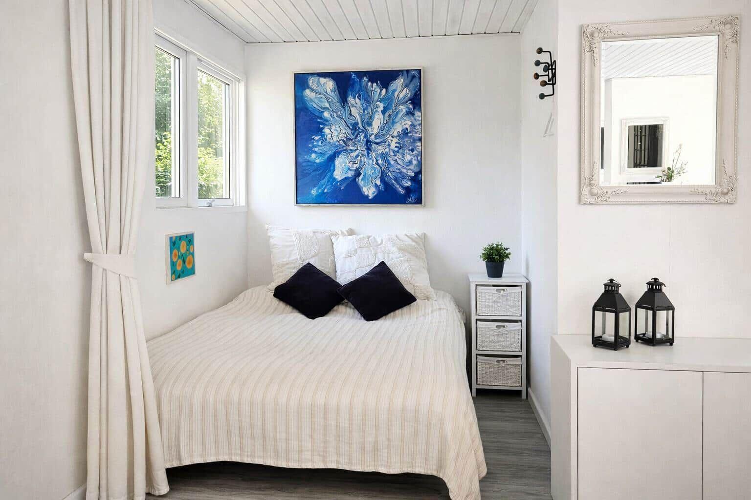 Schlafzimmer mit Bett, Wandbild und Spiegel. Neben dem Bett steht ein Nachttisch mit Körben.