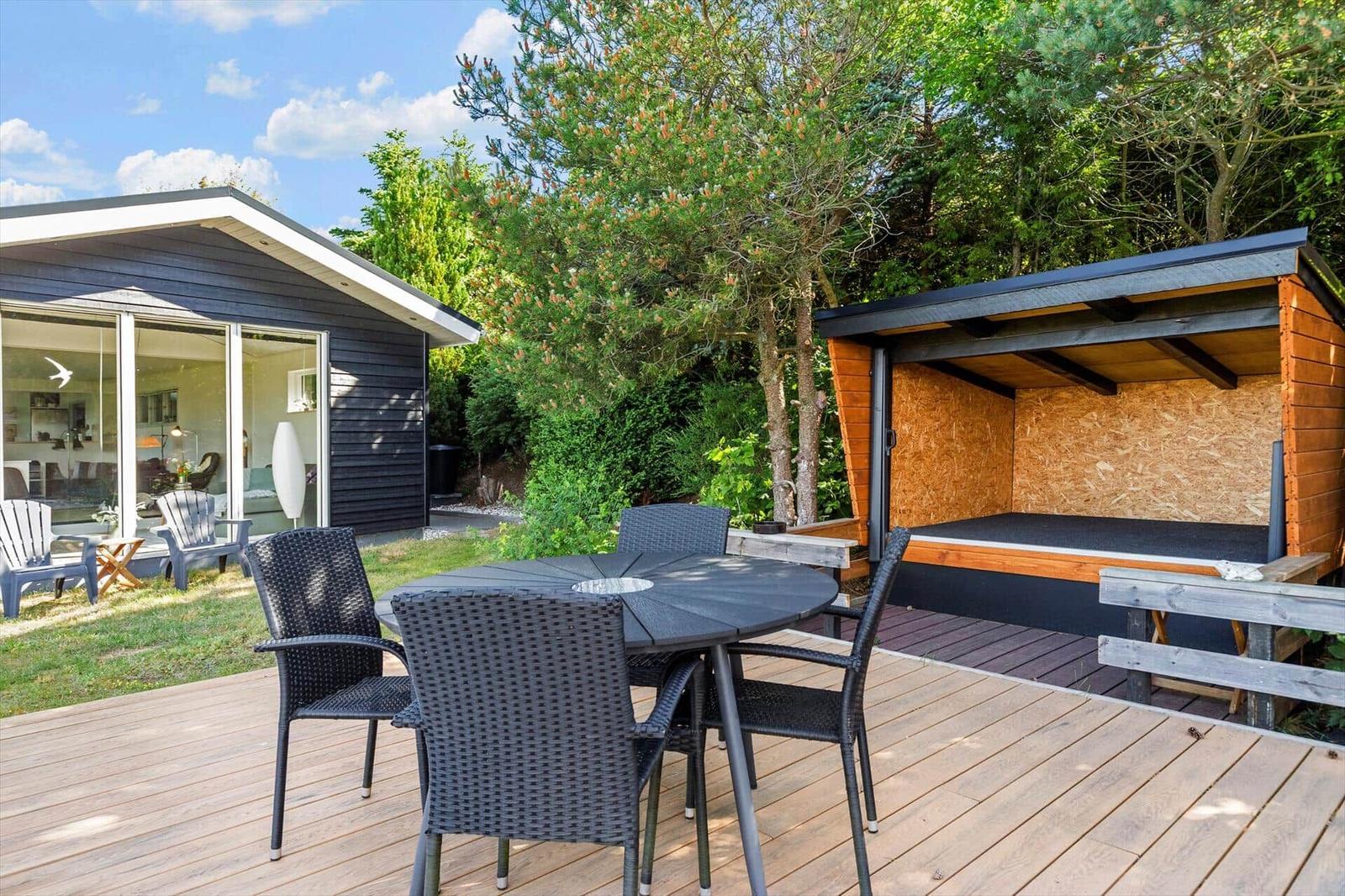 Terrasse mit Tisch und Stühlen, Holzterrasse, Grillhütte und Wohnhaus mit Glasfront.
