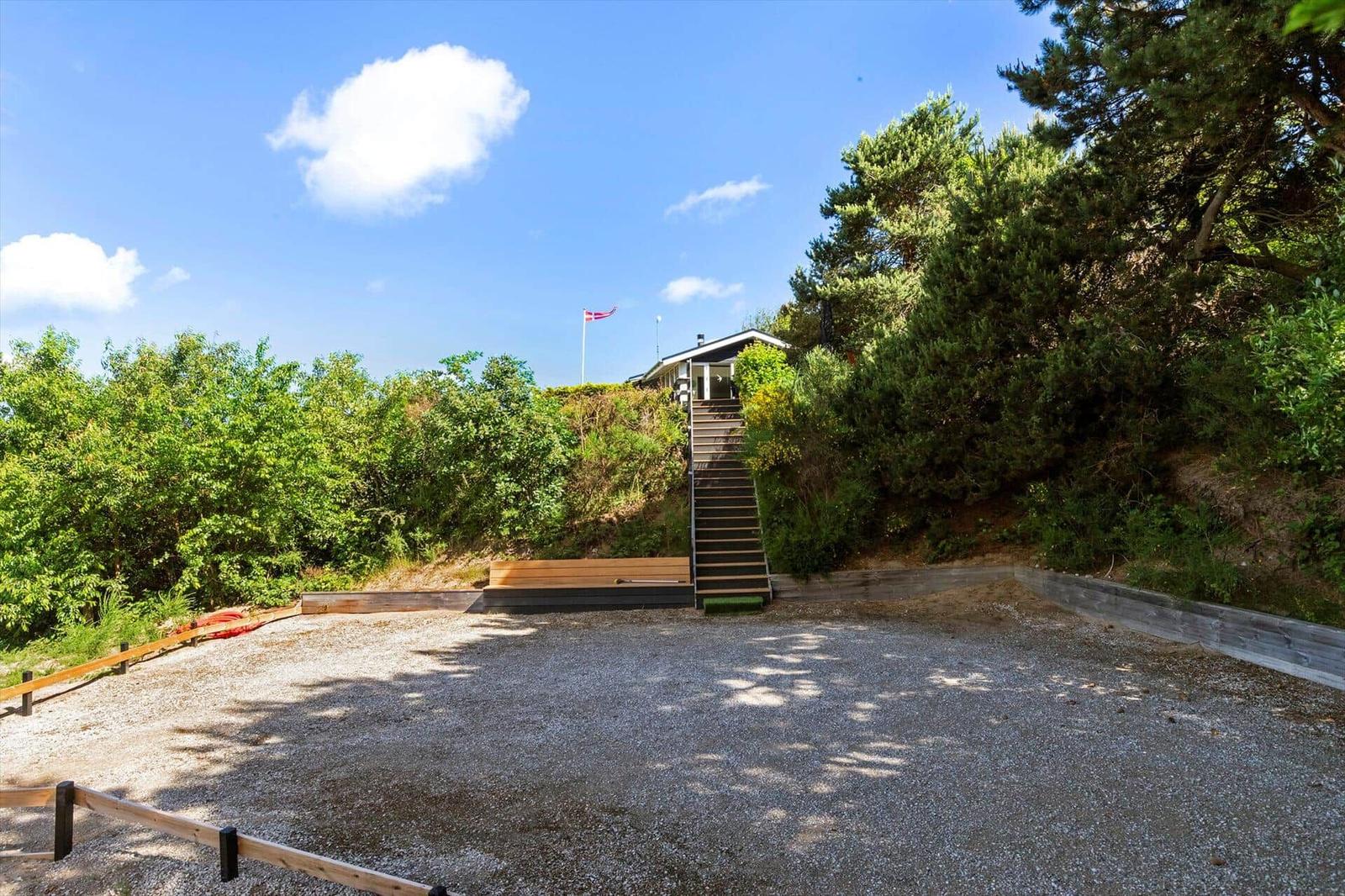 Haus auf Hügel mit Treppe, umgeben von Bäumen und Gras. Gravelfläche im Vordergrund.
