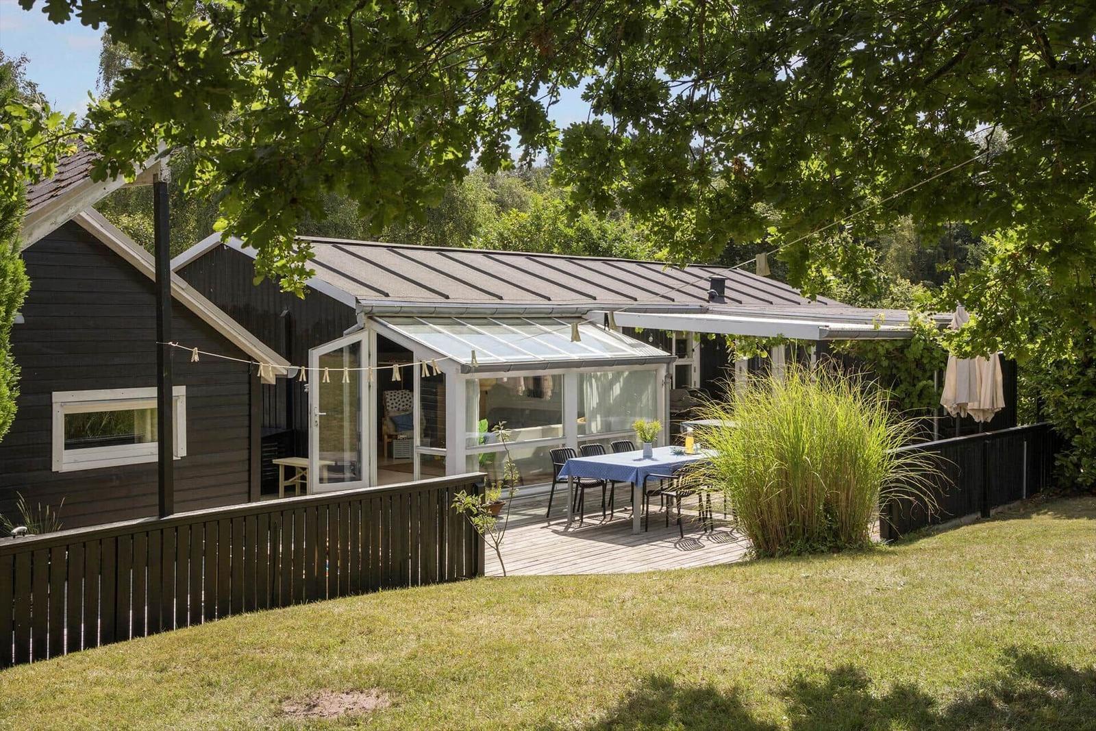 Haus mit Terrasse, Tisch und Stühlen unter Bäumen. Grünfläche und Schutzdach.
