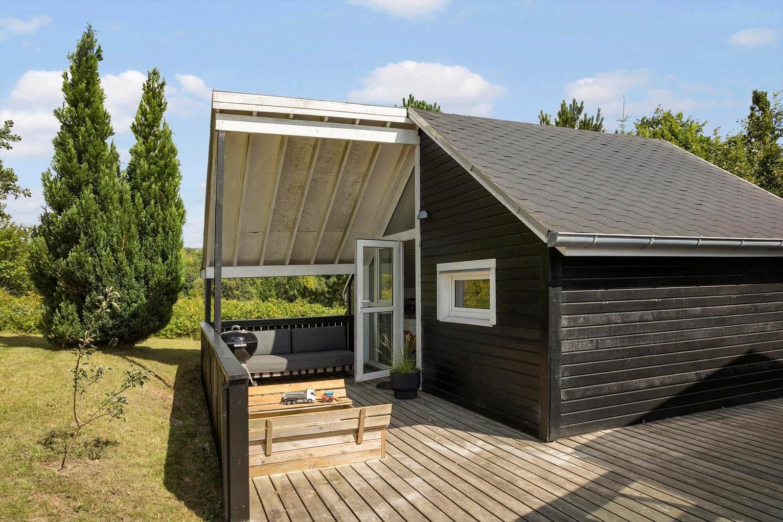 Schwarzes Holzhaus mit Terrasse, Sitzbank und Grill unter Dach.