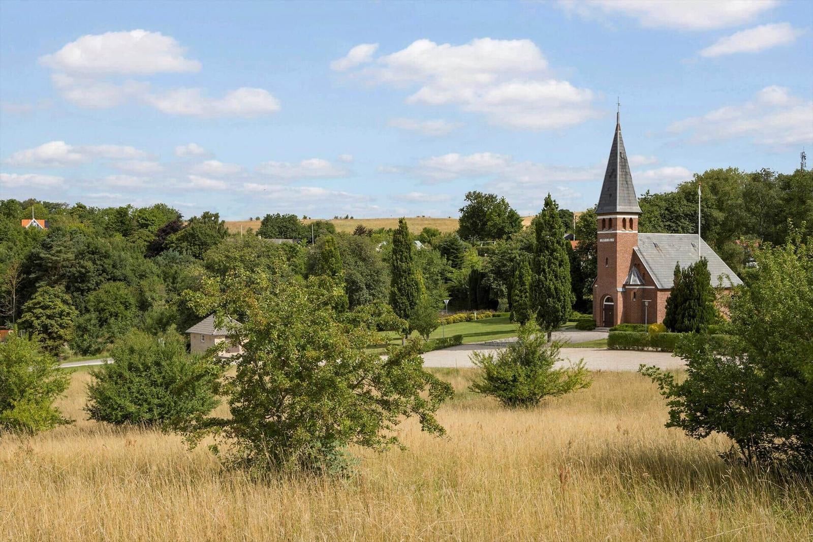 Rotes Kirchengebäude mit Turm, umgeben von Grünflächen und Bäumen unter blauem Himmel.