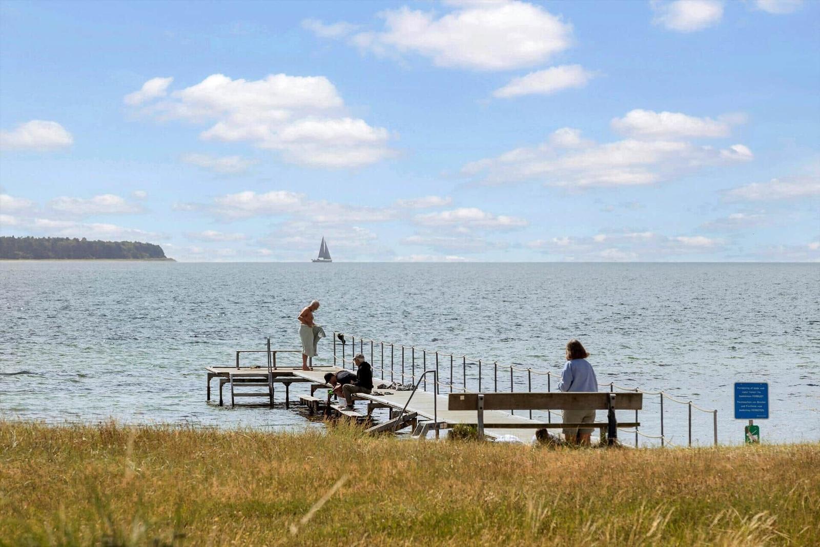 Ein Steg führt ins Wasser, wo Personen stehen und sitzen. Ein Segelboot ist im Hintergrund zu sehen.