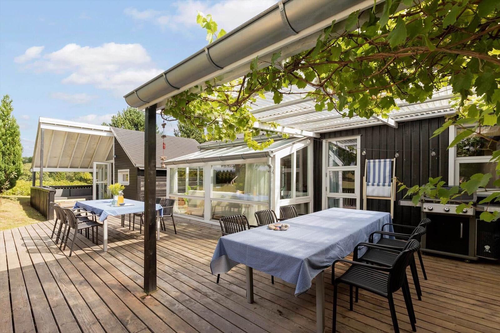 Holzterrasse mit Tischen, Stühlen und Grill unter Kletterpflanzen.