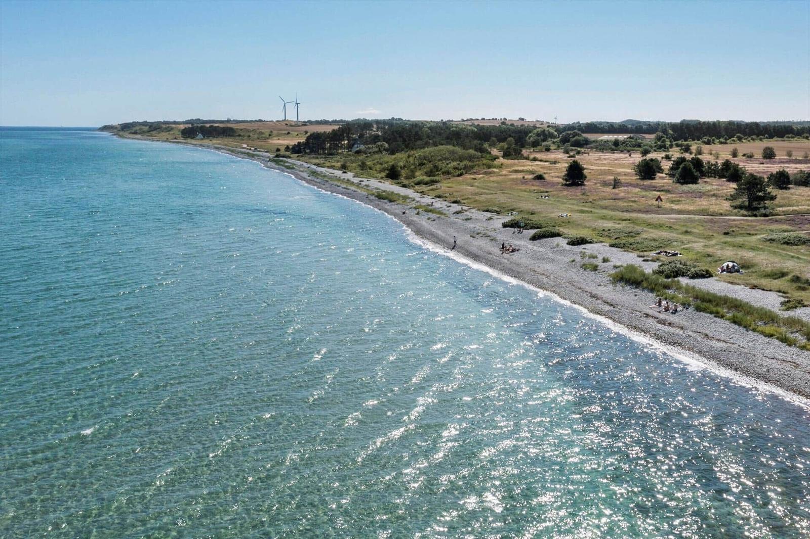 Strand mit grauem Kies, grüne Landschaft und Windkraftanlagen im Hintergrund.