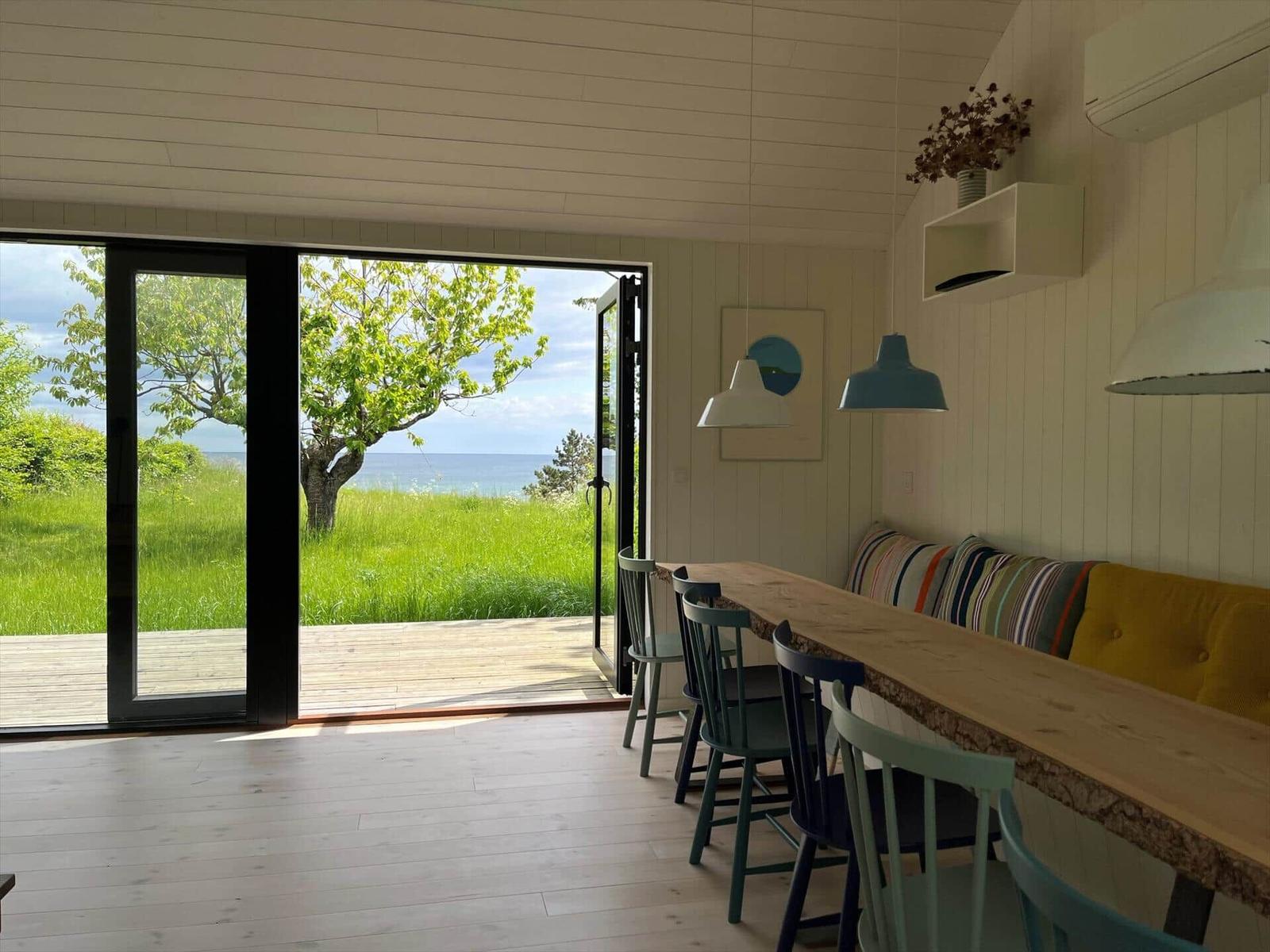Essbereich mit Holztisch und Stühlen. Durch Glastür Blick auf Grasland und Meer.