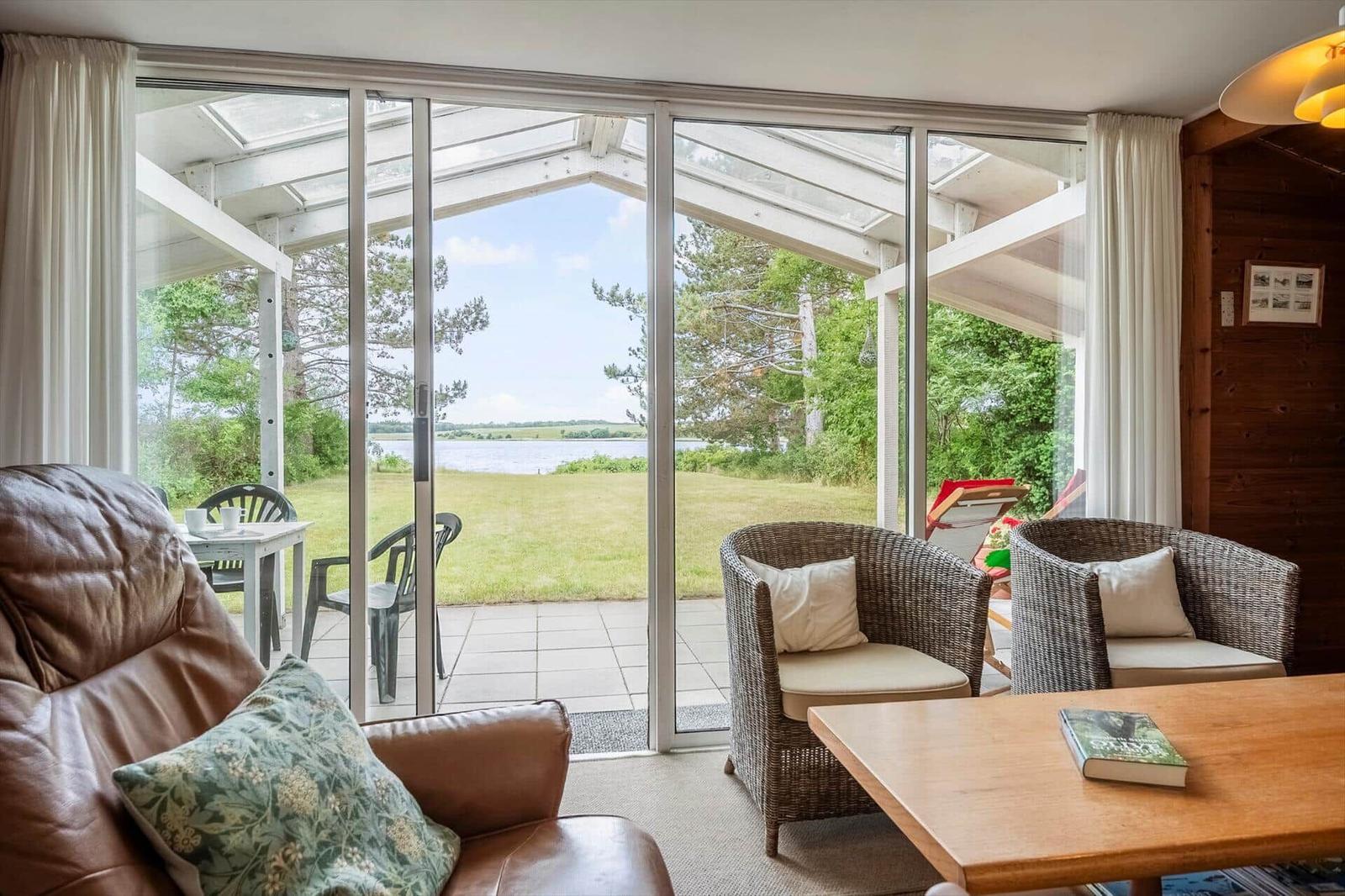 Wohnbereich mit Blick auf einen Garten und See. Rattanstühle, Holztisch und Ledersofa.