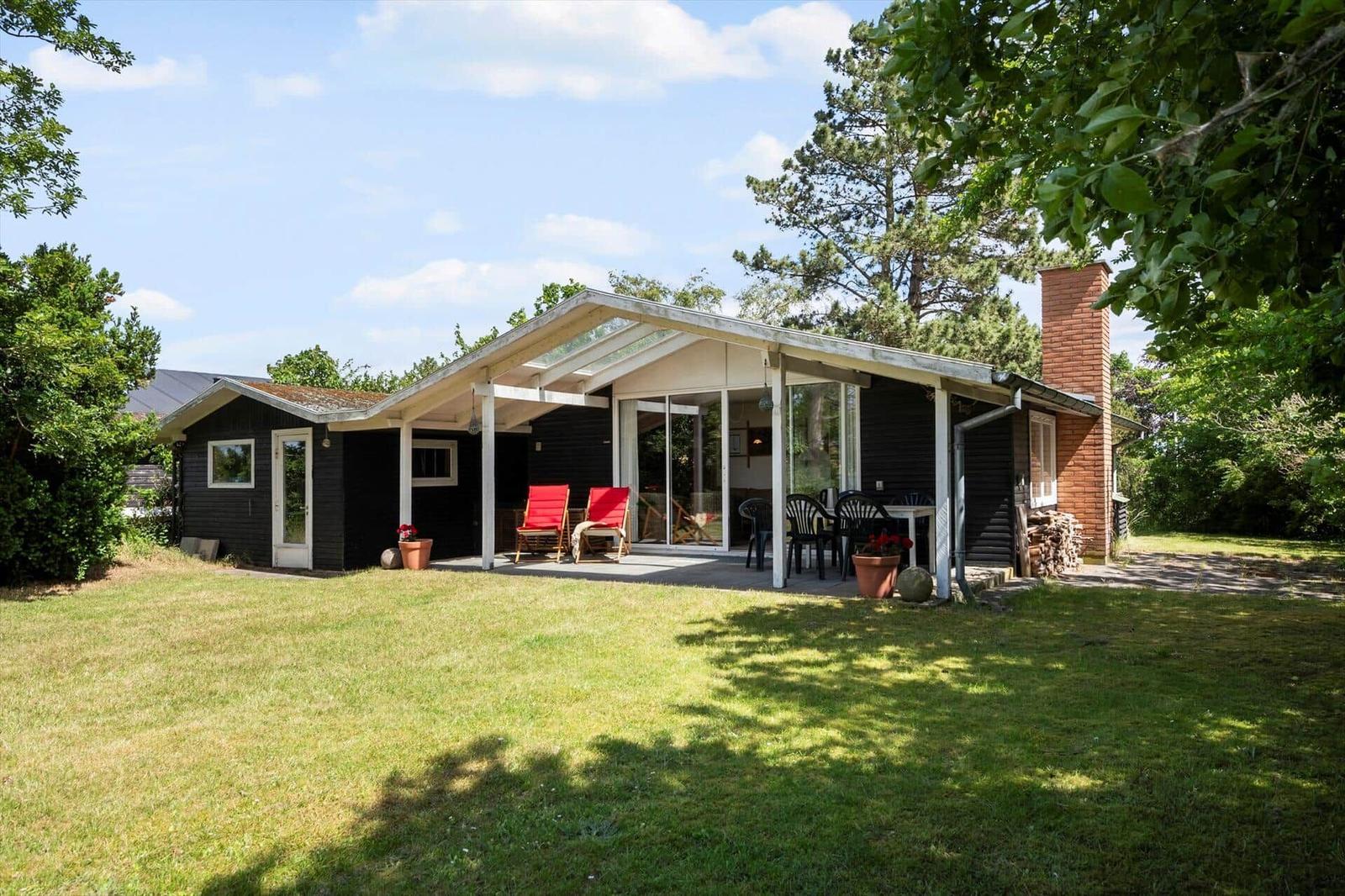 Ein Haus mit Terrasse, roten Stühlen und grünem Garten. Holzhaus mit Kamin und Glasfront.