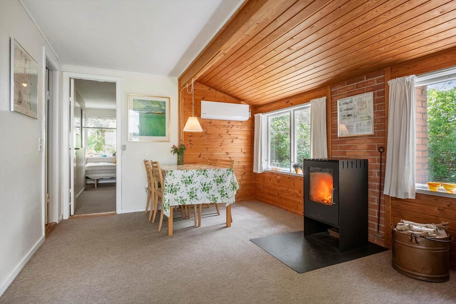 Raum mit Esstisch, Kamin und Blick auf Grün. Holzdecke und Wand. Schlafzimmer im Hintergrund.